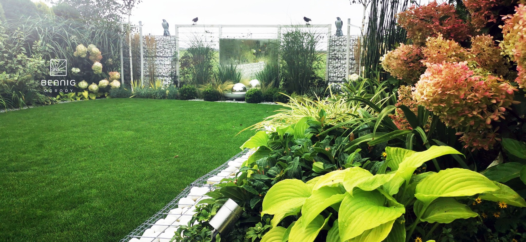 Nowoczesny ogród z równo przystrzyżonym trawnikiem, gabionami wypełnionymi białymi kamieniami i dekoracyjnymi rzeźbami ptaków na ogrodzeniu. Hortensje w odcieniach różu i bieli.