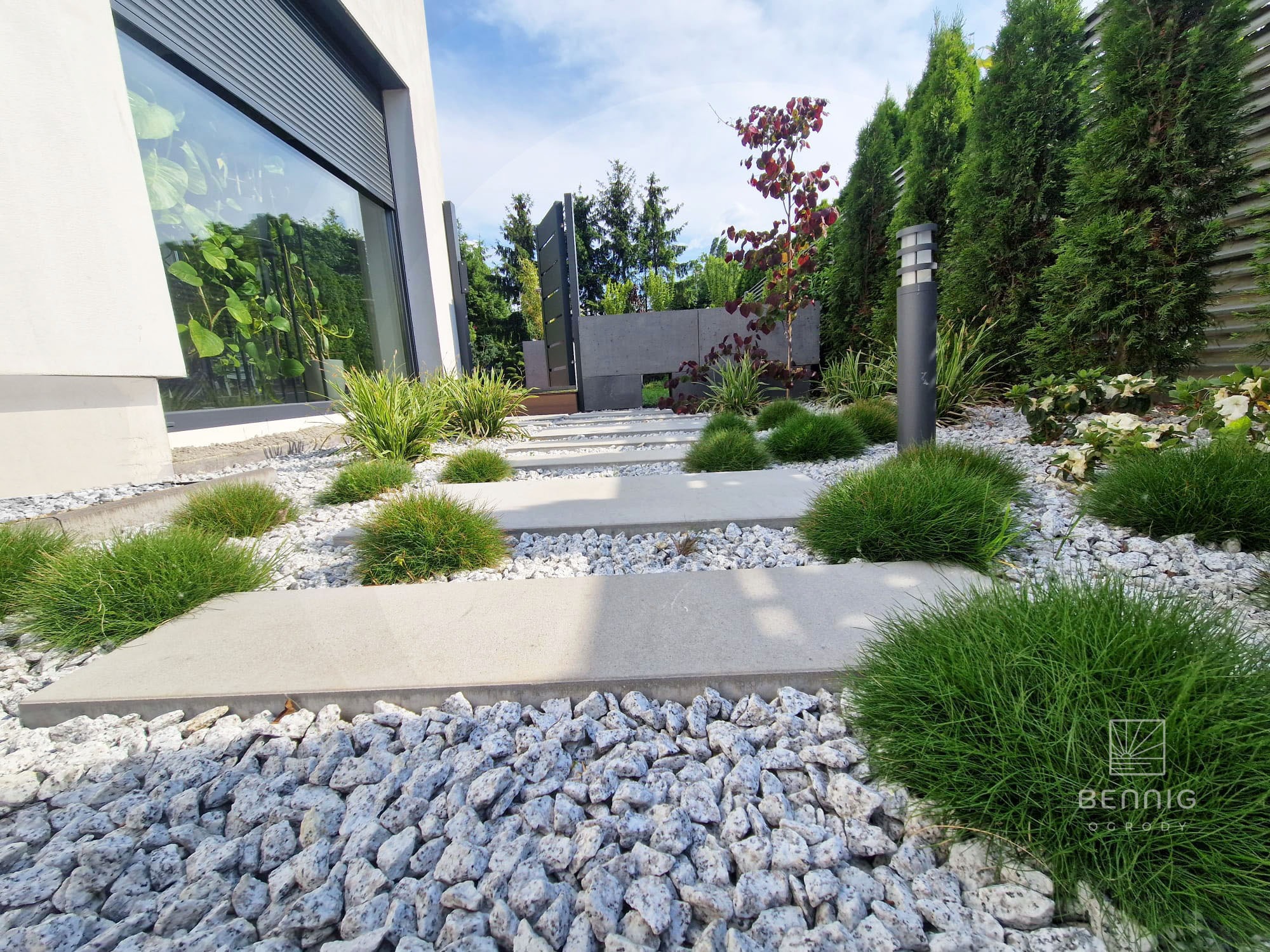 Nowoczesny ogród z białym żwirem, betonowymi płytami ścieżki i ozdobnymi trawami. Minimalistyczny design z lampą ogrodową i drzewem o purpurowych liściach w tle.