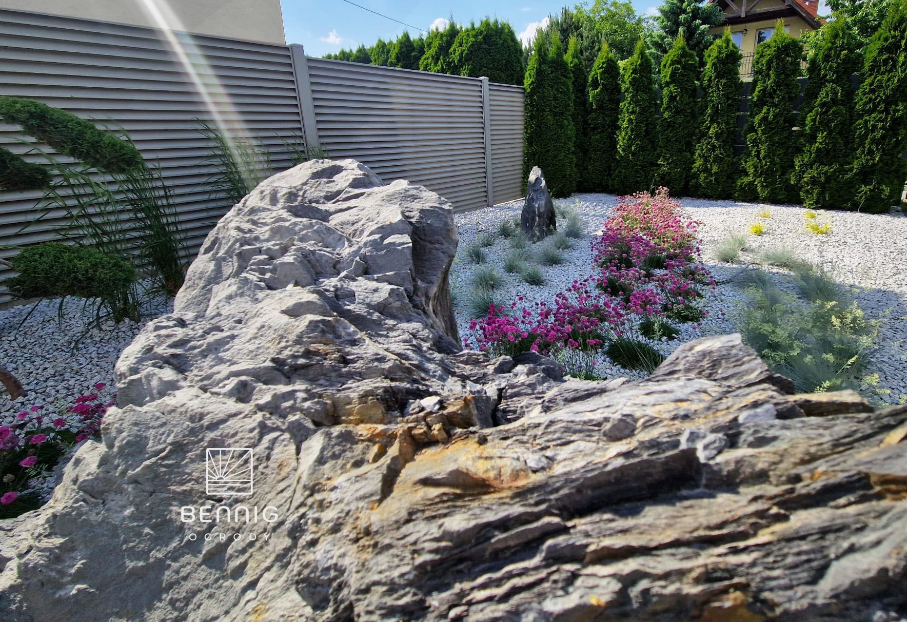 Ogród skalny z dużym, szarym kamieniem na pierwszym planie, białym żwirem, różowymi kwiatami i tujami w tle. Widoczny fragment ogrodzenia i logo firmy.