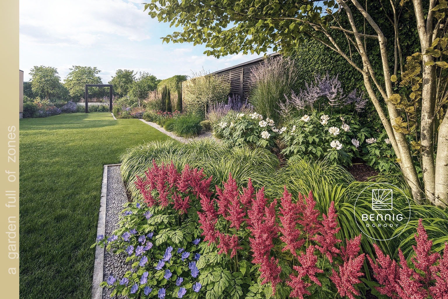 Pełen zieleni, wielopoziomowy ogród z kwitnącymi astilbe, trawami ozdobnymi i drzewami. W tle pergola i zadbany trawnik. Widoczny fragment logo firmy.