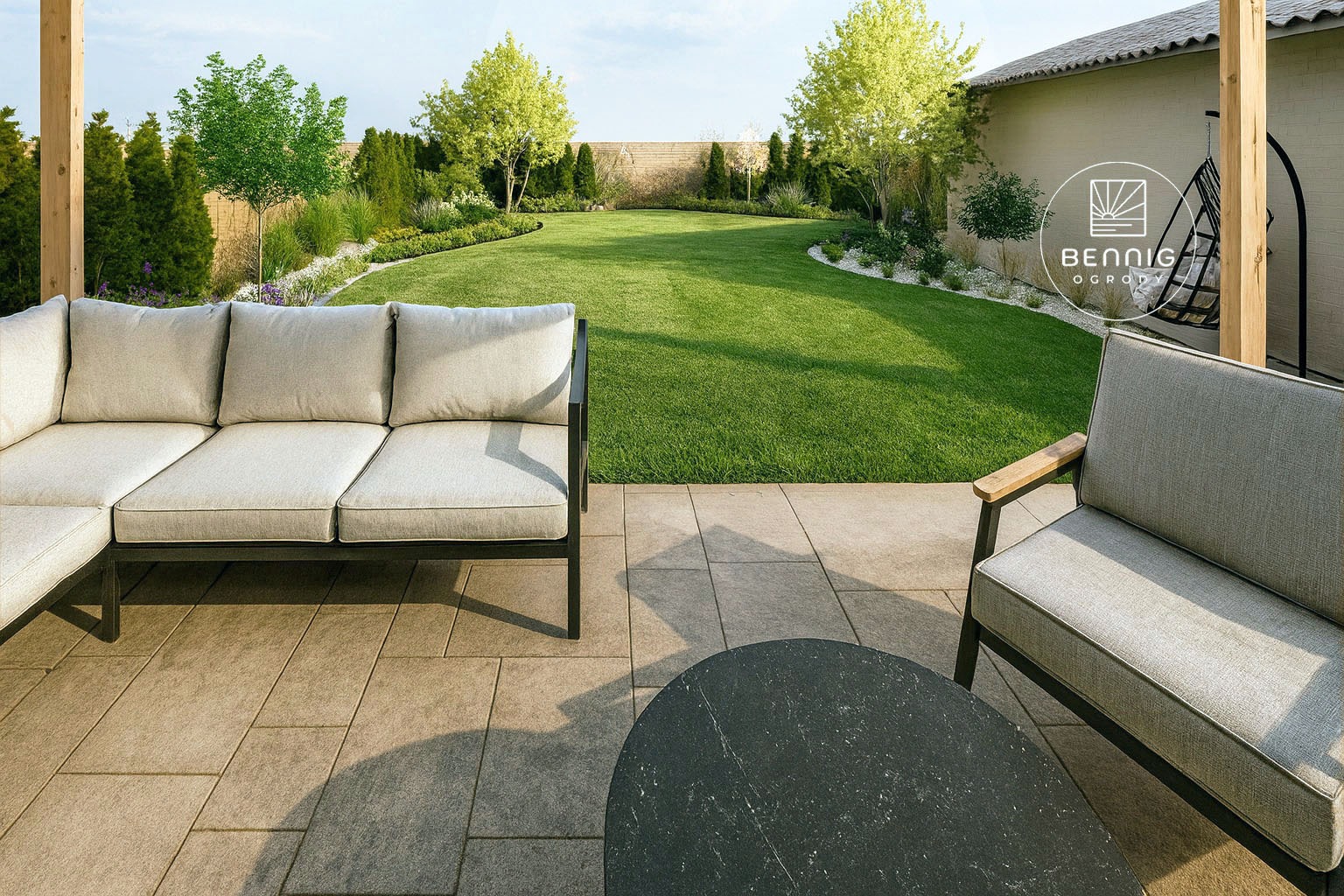 Nowoczesny ogród z wypielęgnowanym trawnikiem, meblami ogrodowymi i dekoracyjnymi nasadzeniami. Widoczny narożnik i fotel na kamiennej posadzce.