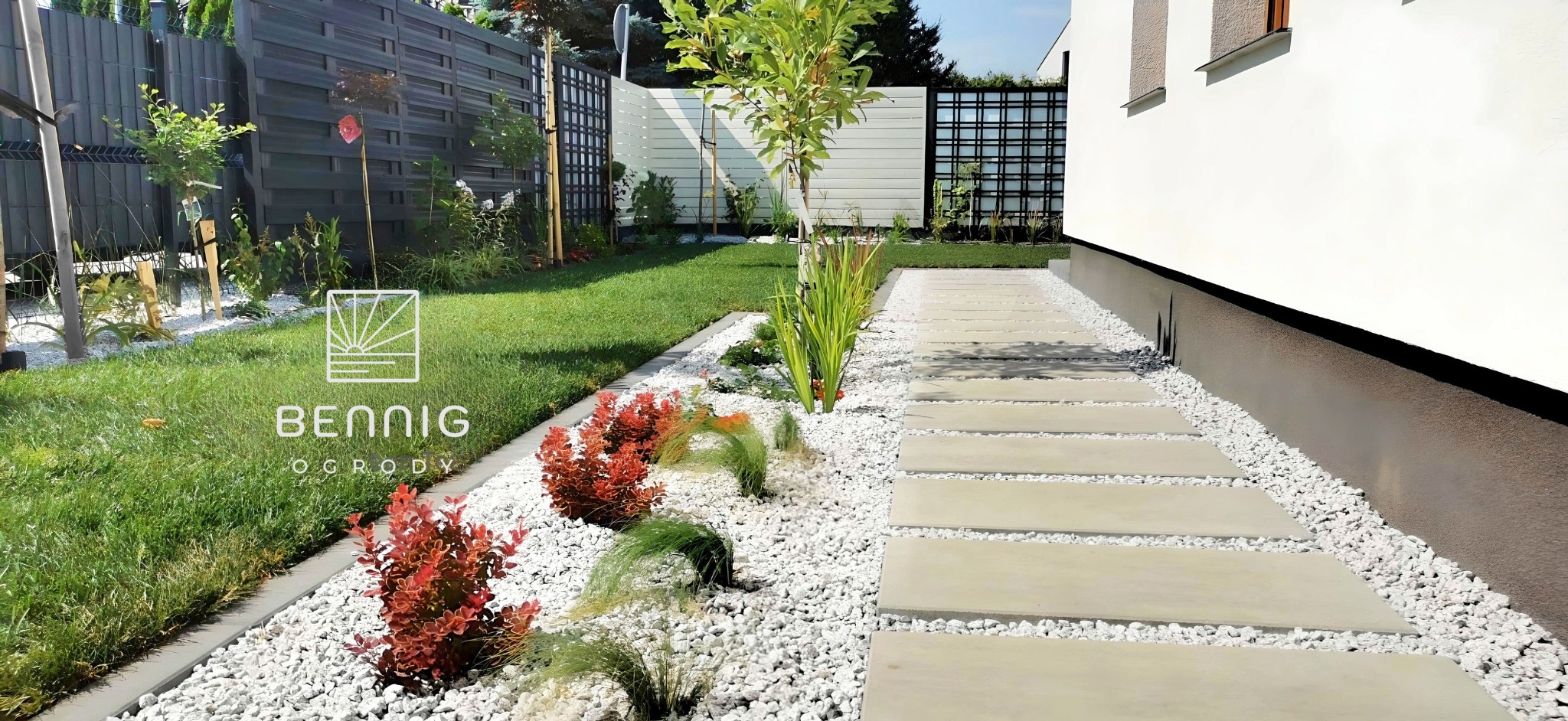 Nowoczesny ogród z białymi kamieniami, ścieżką z płyt i ozdobnymi roślinami. Widoczny fragment budynku i ogrodzenie. Minimalistyczny design.