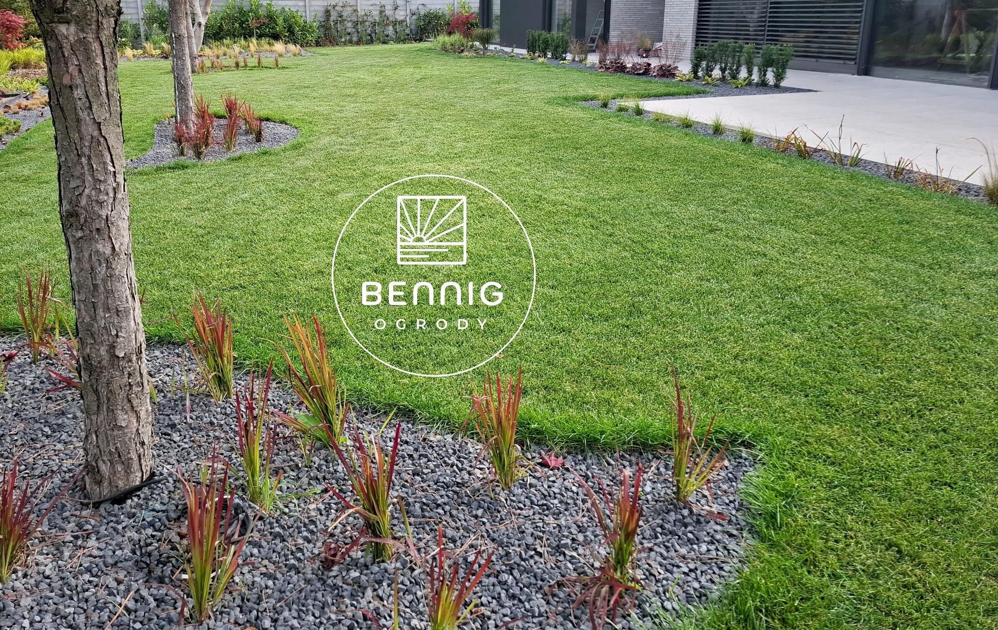Zadbany trawnik z nasadzeniami ozdobnymi i kamieniami, widok z poziomu gruntu. W tle nowoczesny budynek z szarymi elementami. Logo firmy na pierwszym planie.