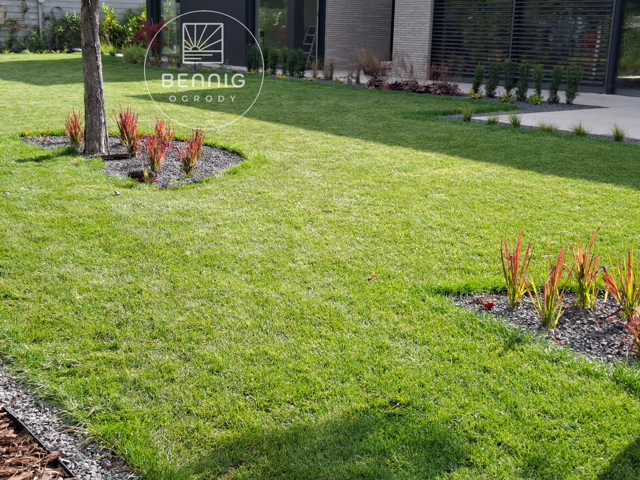 Zadbany trawnik z rabatami wysypanymi grysem, obrzeża z kory, nowoczesny dom w tle. Widok na równo skoszoną trawę i dekoracyjne nasadzenia.