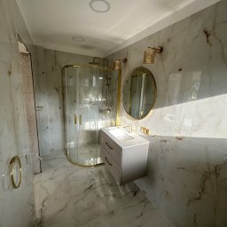 Perfect-Dom Piotr Dombrowski - Wykończona łazienka z kabiną prysznicową w złotej ramie, umywalką z szafką i okrągłym lustrem na ścianie wyłożonej marmurowymi płytkami.