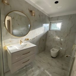 Perfect-Dom Piotr Dombrowski - Wykończona łazienka z marmurowymi płytkami, okrągłym lustrem w złotej ramie i beżową szafką pod umywalką. Widoczne oświetlenie i toaleta. Promienie słoneczne wpadają przez okno.