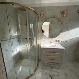 Perfect-Dom Piotr Dombrowski - Wykończona łazienka z kabiną prysznicową w złotej ramie, okrągłym lustrem i umywalką z szafką w marmurowym wykończeniu. Nowoczesny design, jasne płytki.