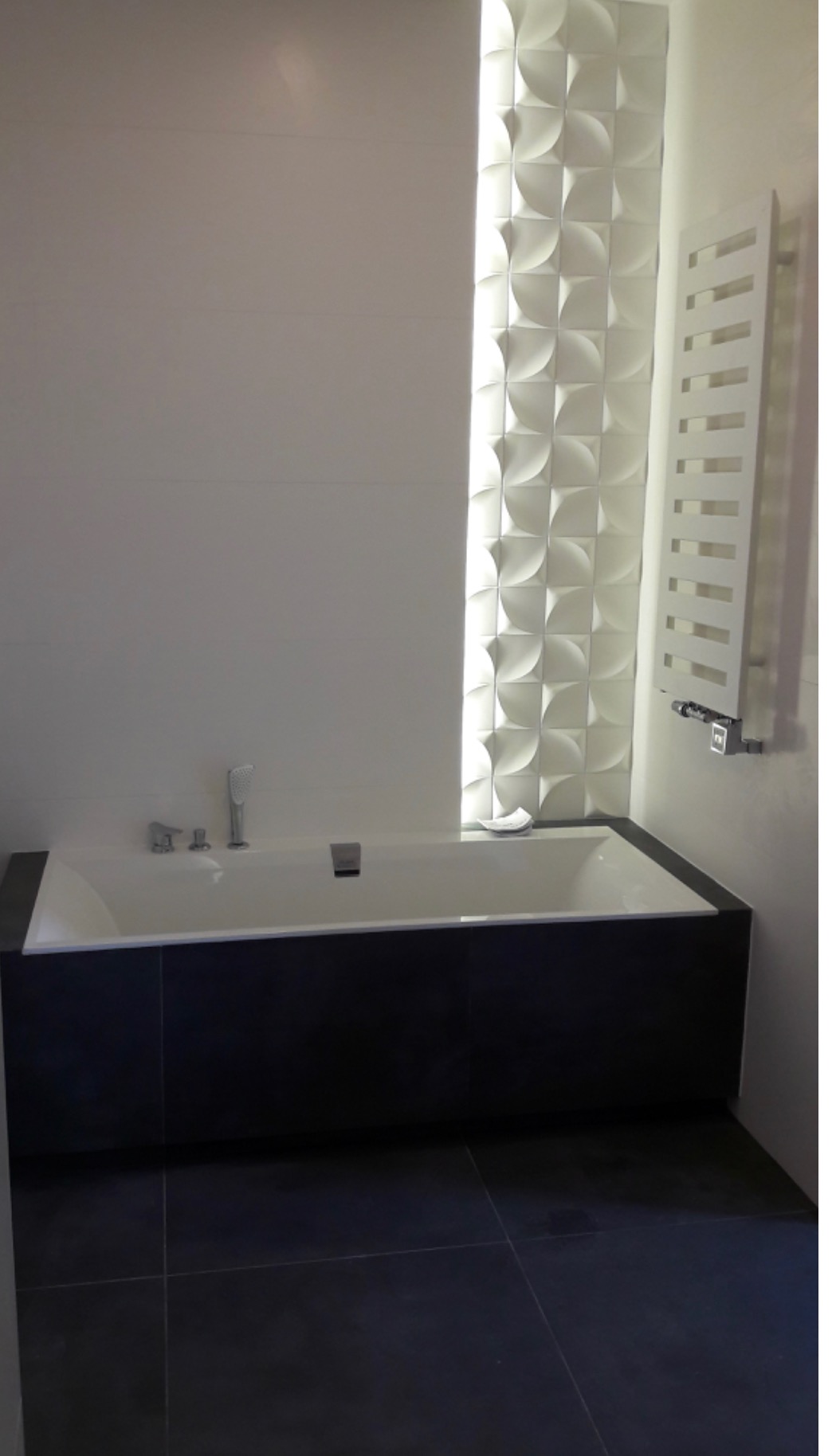 Nowoczesna łazienka z białą wanną obudowaną czarnymi płytkami, dekoracyjną ścianą 3D z podświetleniem i białym grzejnikiem drabinkowym.