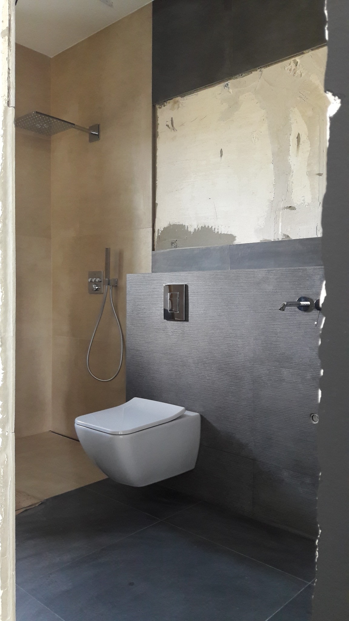 Wnętrze łazienki w trakcie remontu, widoczny podwieszany sedes, szara glazura z poziomym wzorem, deszczownica, ściana przygotowana do położenia gładzi.