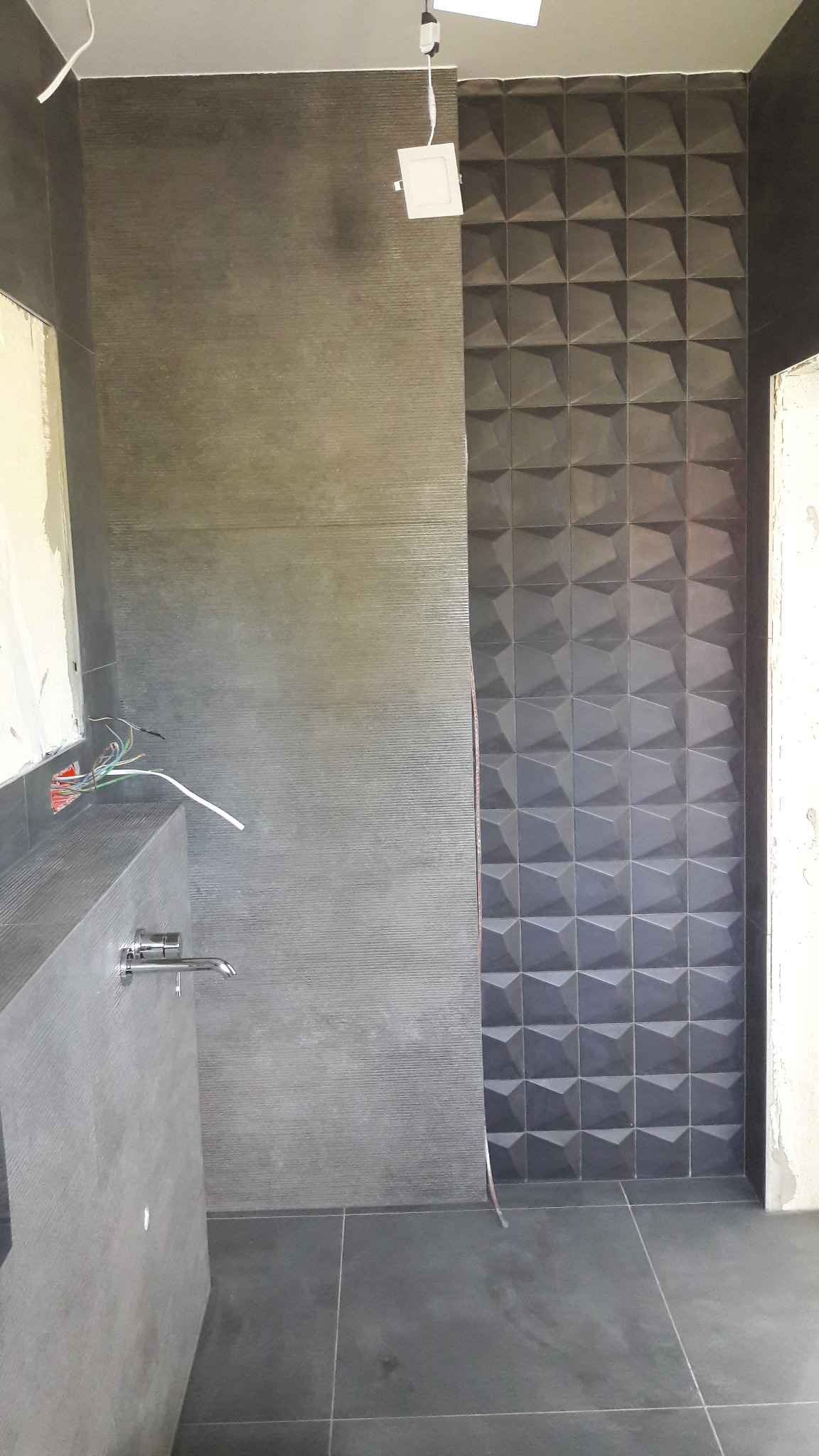 Niedokończona łazienka z szarą, teksturowaną ścianą, trójwymiarowymi płytkami i odsłoniętymi przewodami elektrycznymi nad umywalką z chromowaną baterią.