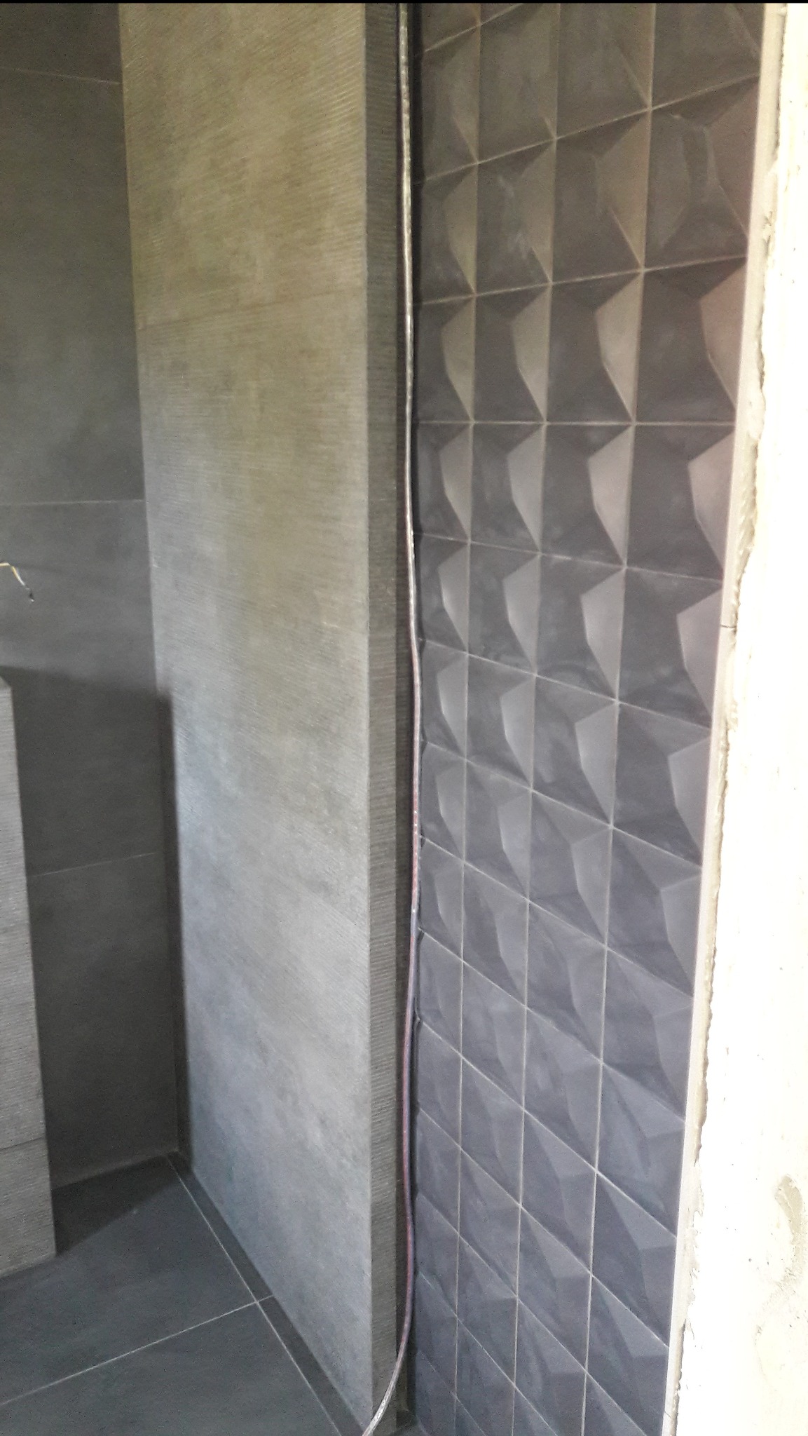 Fragment ściany łazienki z szarą, trójwymiarową glazurą ułożoną w romby, widoczny przewód elektryczny oraz surowy beton w narożniku.