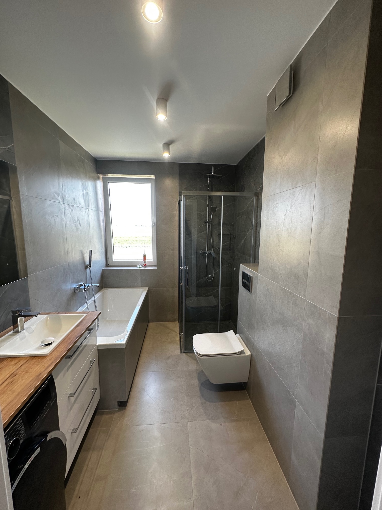 Nowoczesna łazienka w odcieniach szarości z umywalką na drewnianym blacie, wanną przy oknie i kabiną prysznicową typu walk-in.