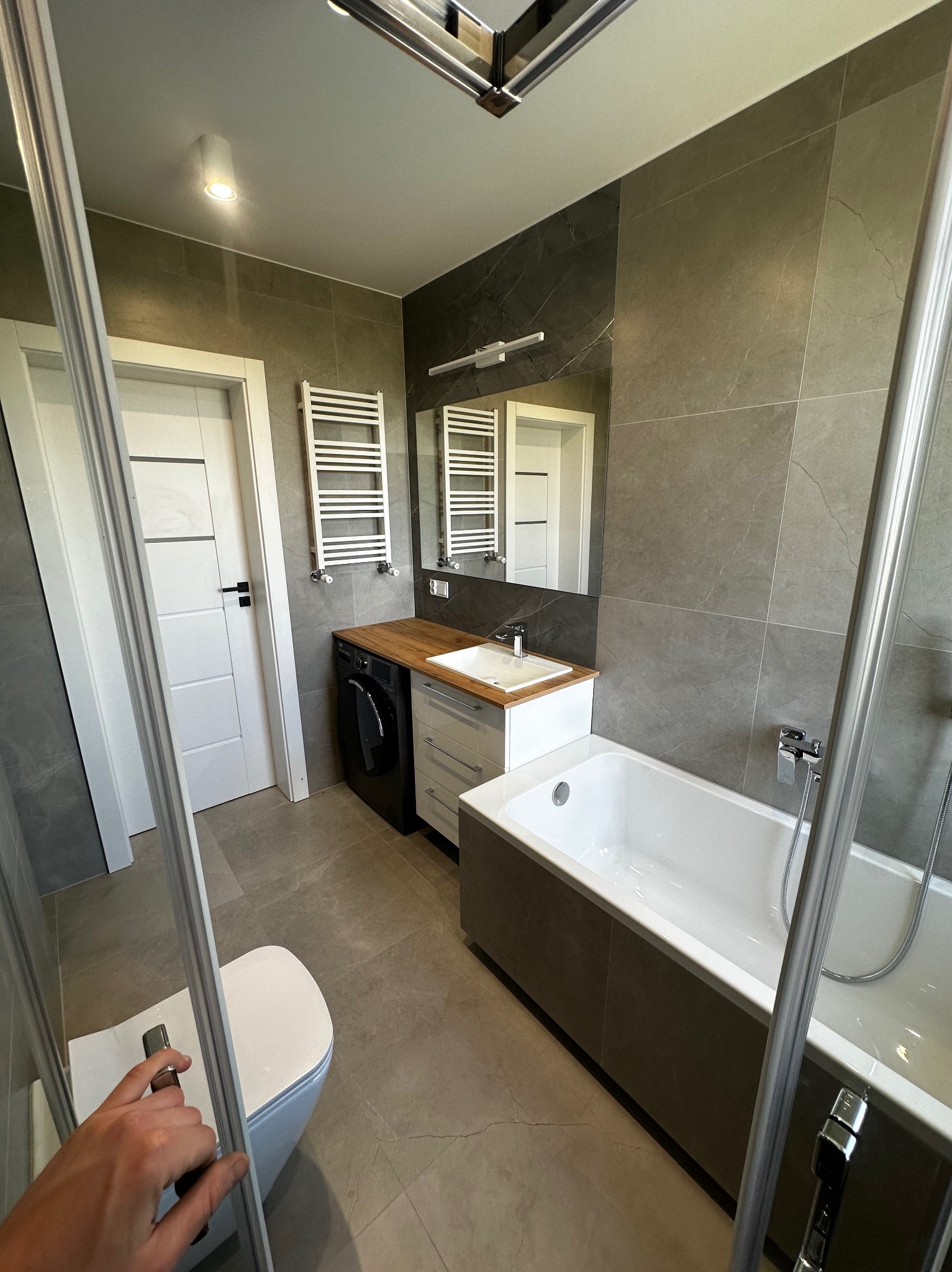 Nowoczesna łazienka z szarą glazurą, białą wanną, umywalką z drewnianym blatem, pralką w zabudowie i grzejnikiem drabinkowym, widziana przez częściowo otwarte szklane drzwi kabiny prysznicowej.