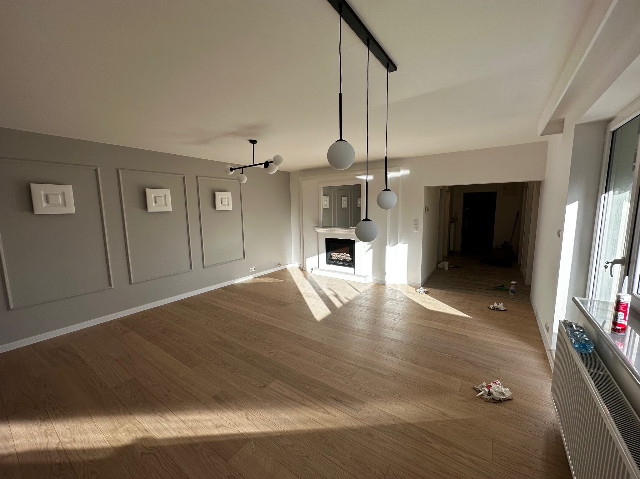 Jasne, przestronne wnętrze po remoncie: parkiet z naturalnego drewna, szara ściana z dekoracyjnymi panelami, nowoczesny kominek i designerskie lampy sufitowe.