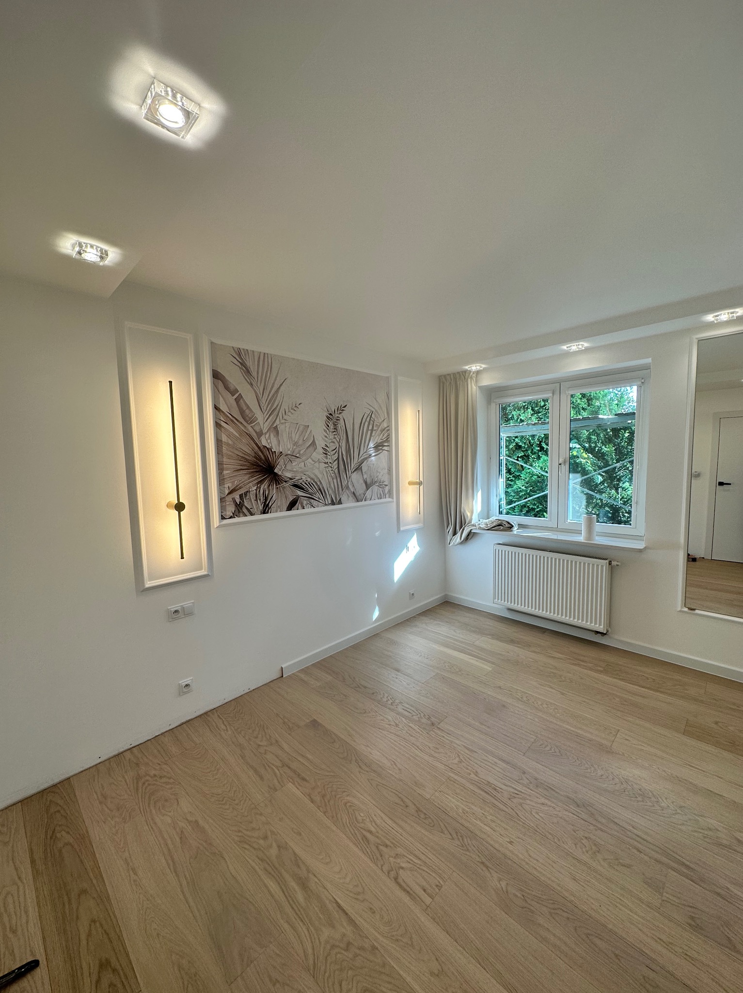 Jasne, przestronne wnętrze pokoju z drewnianą podłogą, białymi ścianami zdobionymi geometrycznymi lampami i graficznym motywem roślinnym, okno z widokiem na zieleń, grzejnik pod oknem...