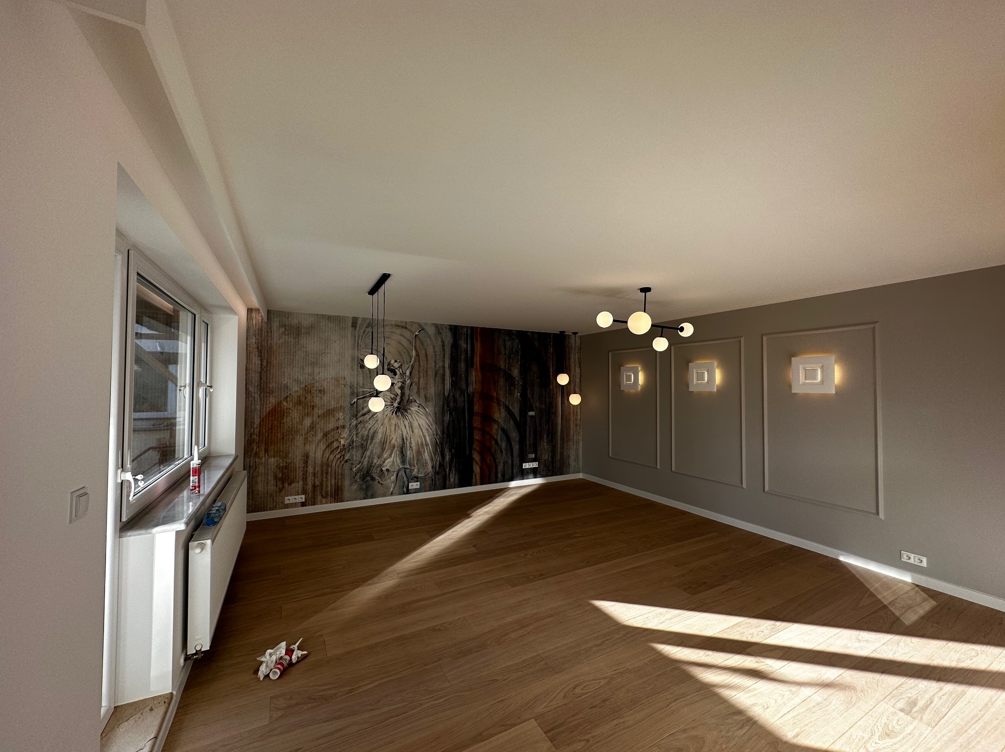 Jasne, przestronne wnętrze z drewnianą podłogą, dekoracyjną tapetą z motywem abstrakcyjnym, nowoczesnym oświetleniem i listwami ściennymi, częściowo wykończone, z widocznymi materiałami budowlanymi...