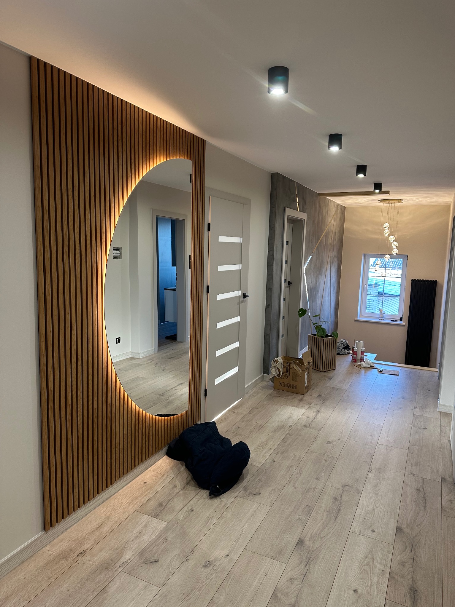 Nowoczesny przedpokój z drewnianą ścianą z lamelami i okrągłym lustrem z podświetleniem LED, jasna podłoga, minimalistyczne oświetlenie sufitowe, widoczne drzwi i dekoracyjna ściana z efektem betonu.