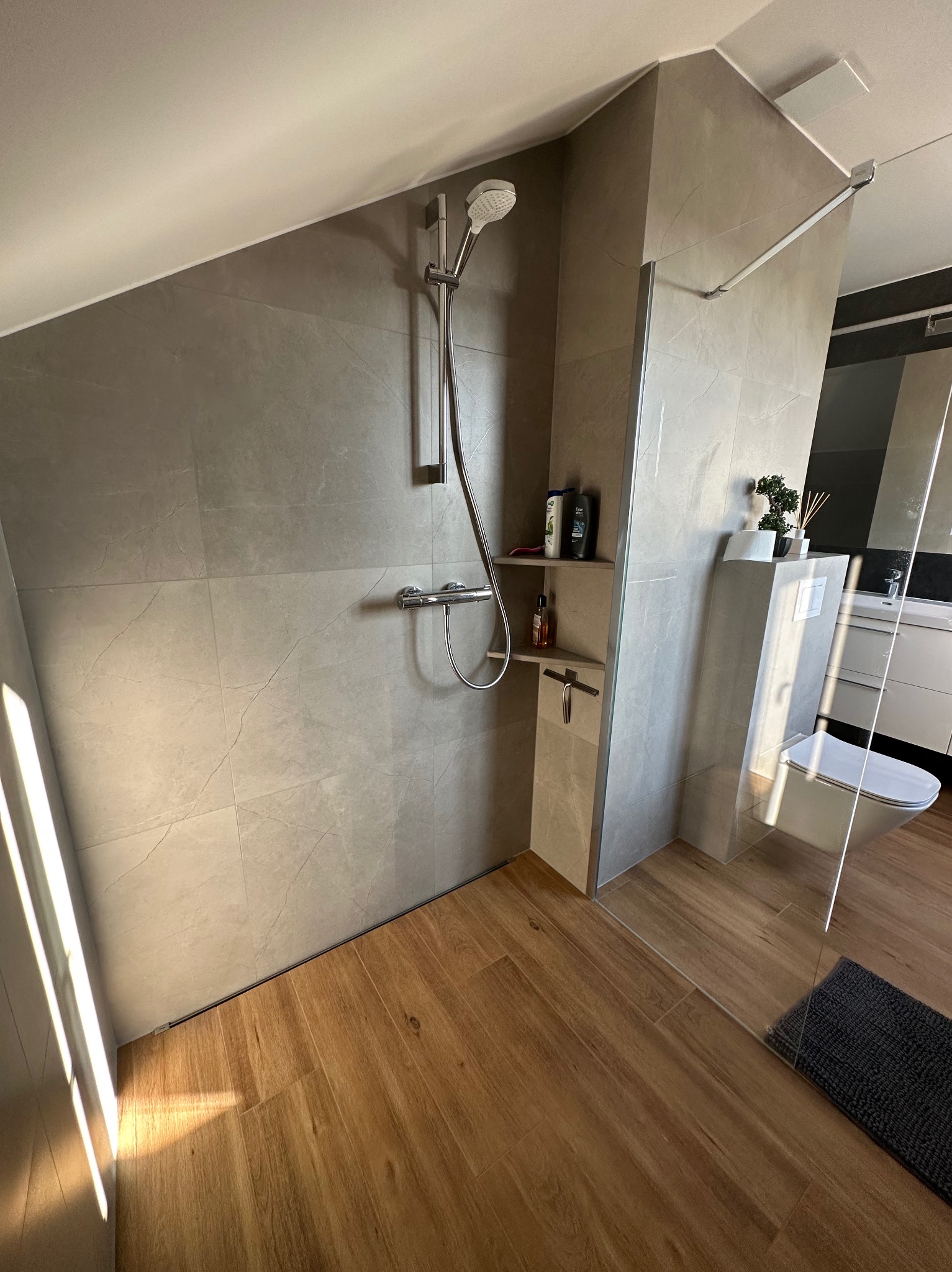 Nowoczesna łazienka z kabiną prysznicową typu walk-in, wykończona szarymi płytkami imitującymi marmur i drewnopodobną podłogą, z wbudowaną półką na kosmetyki i minimalistyczną toaletą.