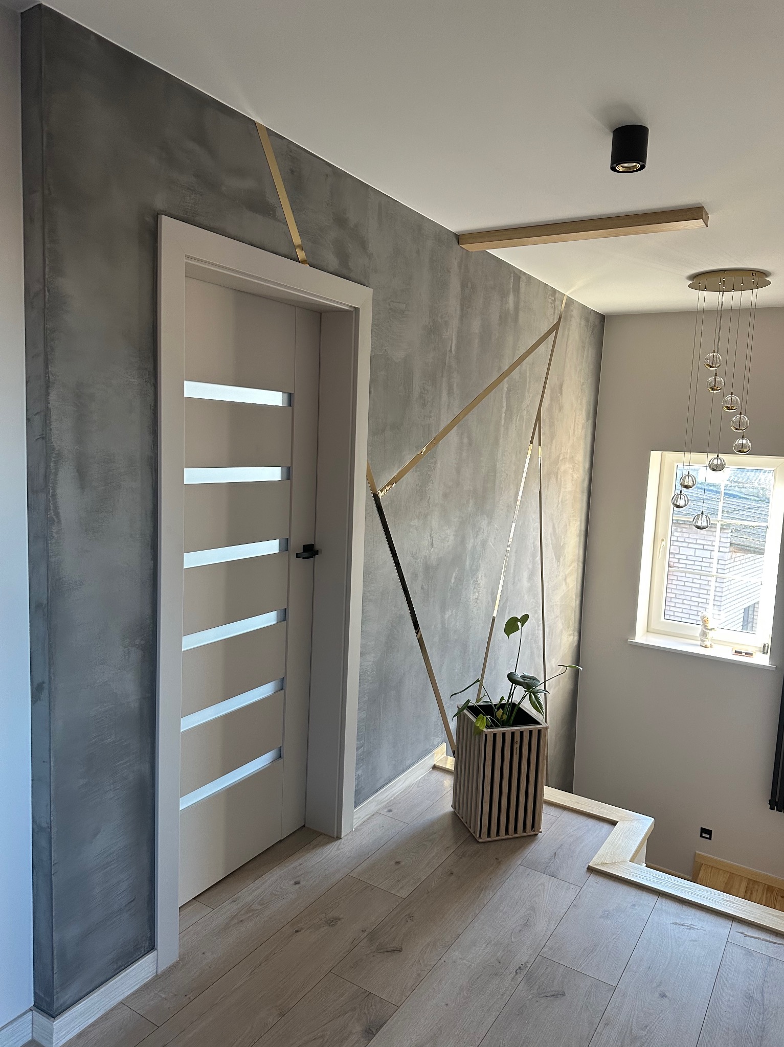 Korytarz z szarą ścianą wykończoną efektem betonu, zdobioną złotymi, geometrycznymi liniami, drewniana podłoga, białe drzwi z przeszkleniami, roślina w drewnianej osłonce, wisząca lampa z kulami...