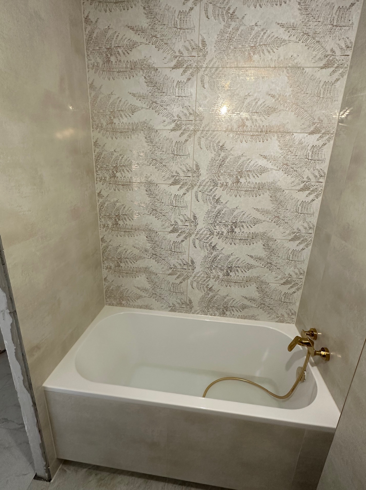 Wykończona łazienka z białą wanną i złotą armaturą. Ściany wyłożone płytkami z motywem roślinnym w odcieniach beżu i brązu. Widoczny fragment niedokończonego obramowania.