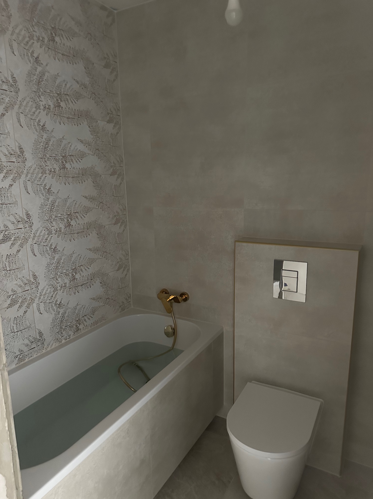 Wykończona łazienka z wanną, toaletą i płytkami w odcieniach szarości. Na ścianie płytki z motywem roślinnym. Bateria wannowa w kolorze złotym.