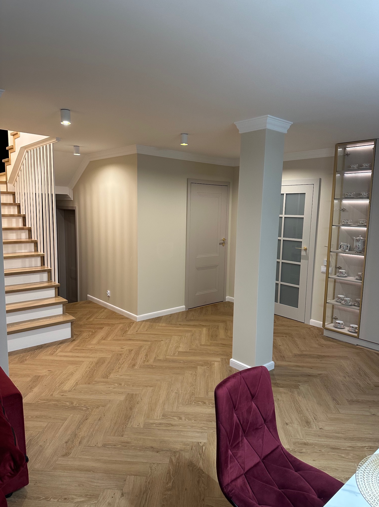 Jasne, przestronne wnętrze z parkietem w jodełkę, schodami z białymi balustradami i kolumną. Widoczne drzwi i przeszklona witryna z dekoracyjną porcelaną.