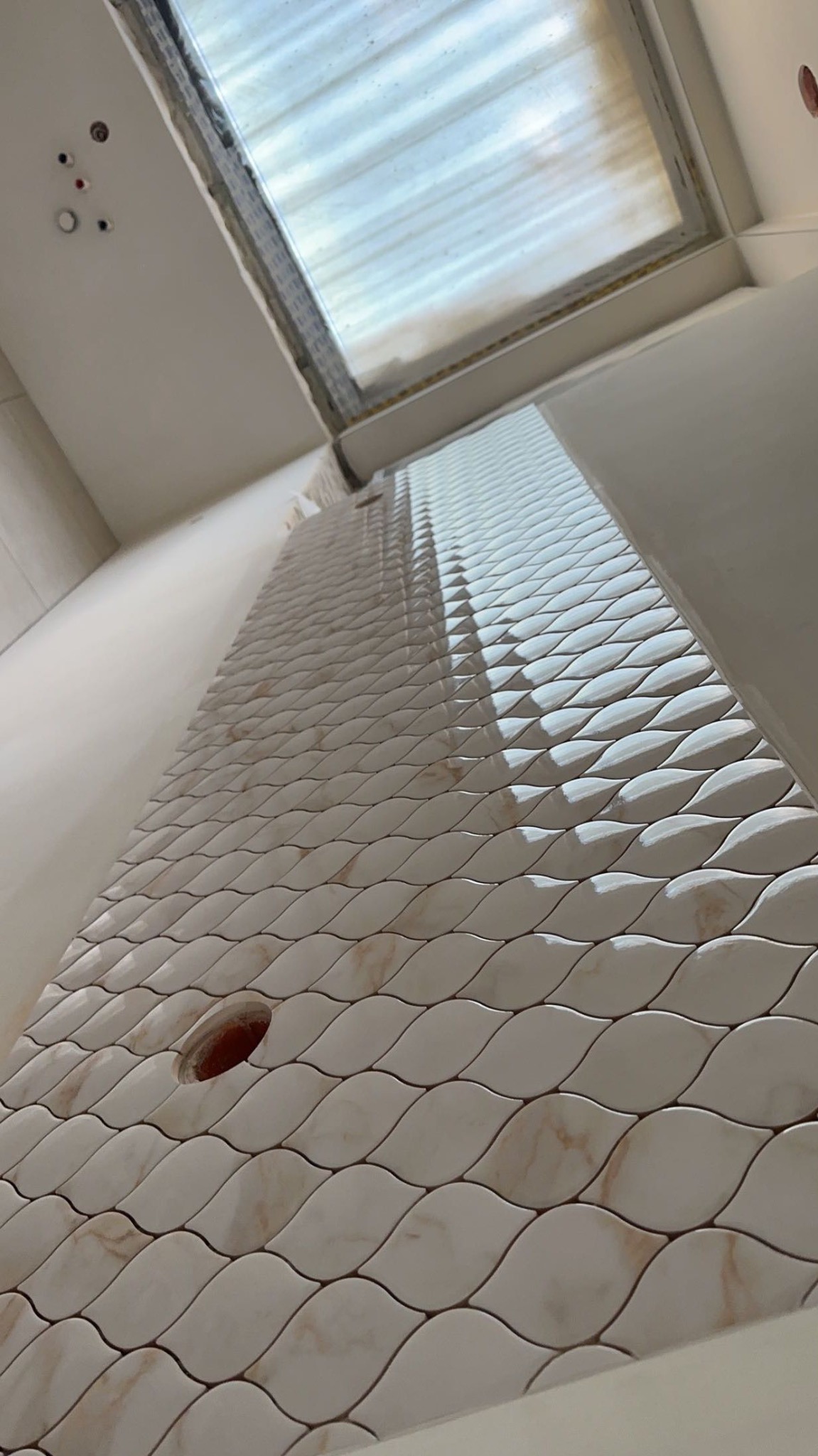 Ukończona ściana z dekoracyjnych, beżowo-białych płytek o falistym wzorze, z otworem na instalację. Widok z dołu, okno w tle.