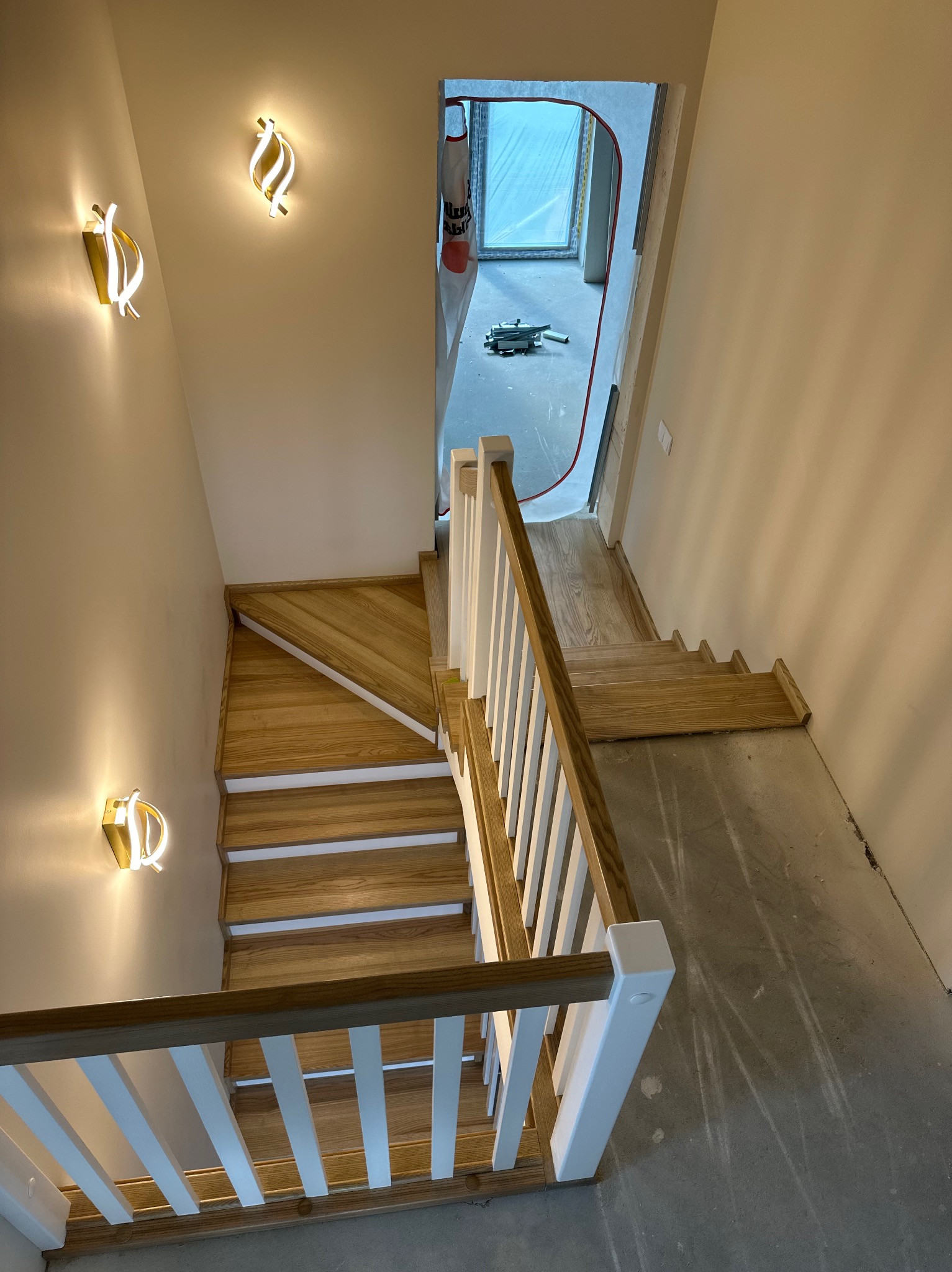 Drewniane schody z białymi balustradami i nowoczesnymi lampami ściennymi. Widok z góry na klatkę schodową w trakcie wykańczania, z widocznym surowym betonem.