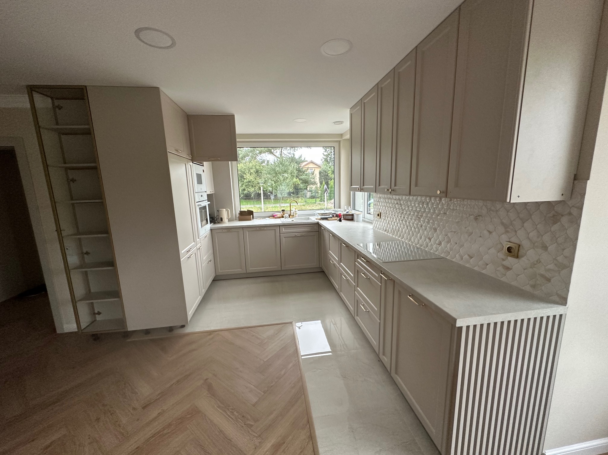 Jasna kuchnia z mozaiką na ścianie i parkietem w jodełkę. Nowoczesne szafki z subtelnymi uchwytami i blatem z szarego kamienia. Duże okno wpuszcza naturalne światło.