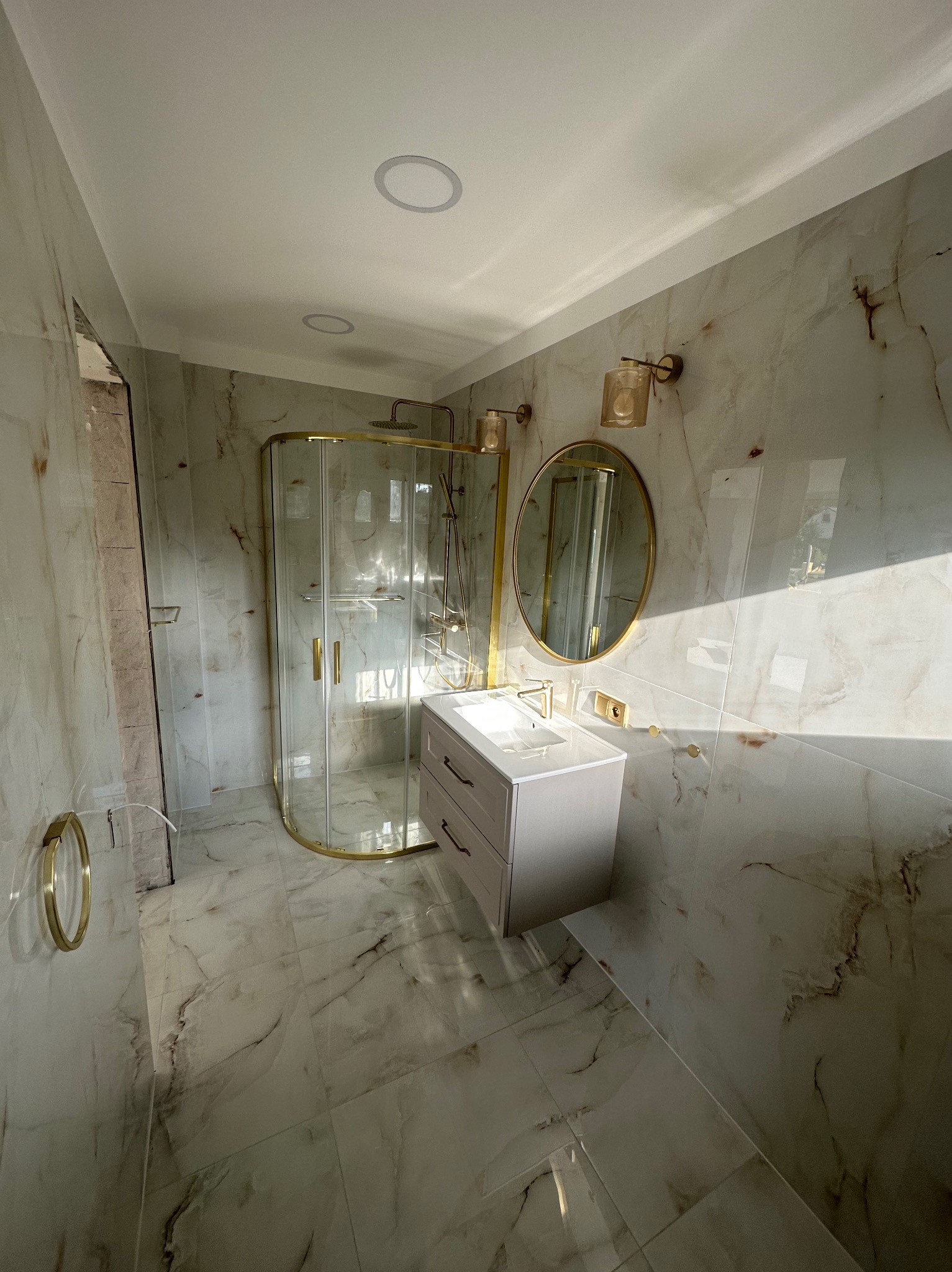 Wykończona łazienka z kabiną prysznicową w złotej ramie, umywalką z szafką i okrągłym lustrem na ścianie wyłożonej marmurowymi płytkami.