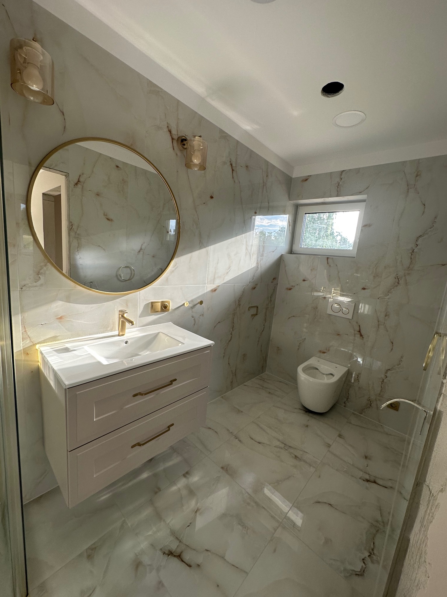 Wykończona łazienka z marmurowymi płytkami, okrągłym lustrem w złotej ramie i beżową szafką pod umywalką. Widoczne oświetlenie i toaleta. Promienie słoneczne wpadają przez okno.