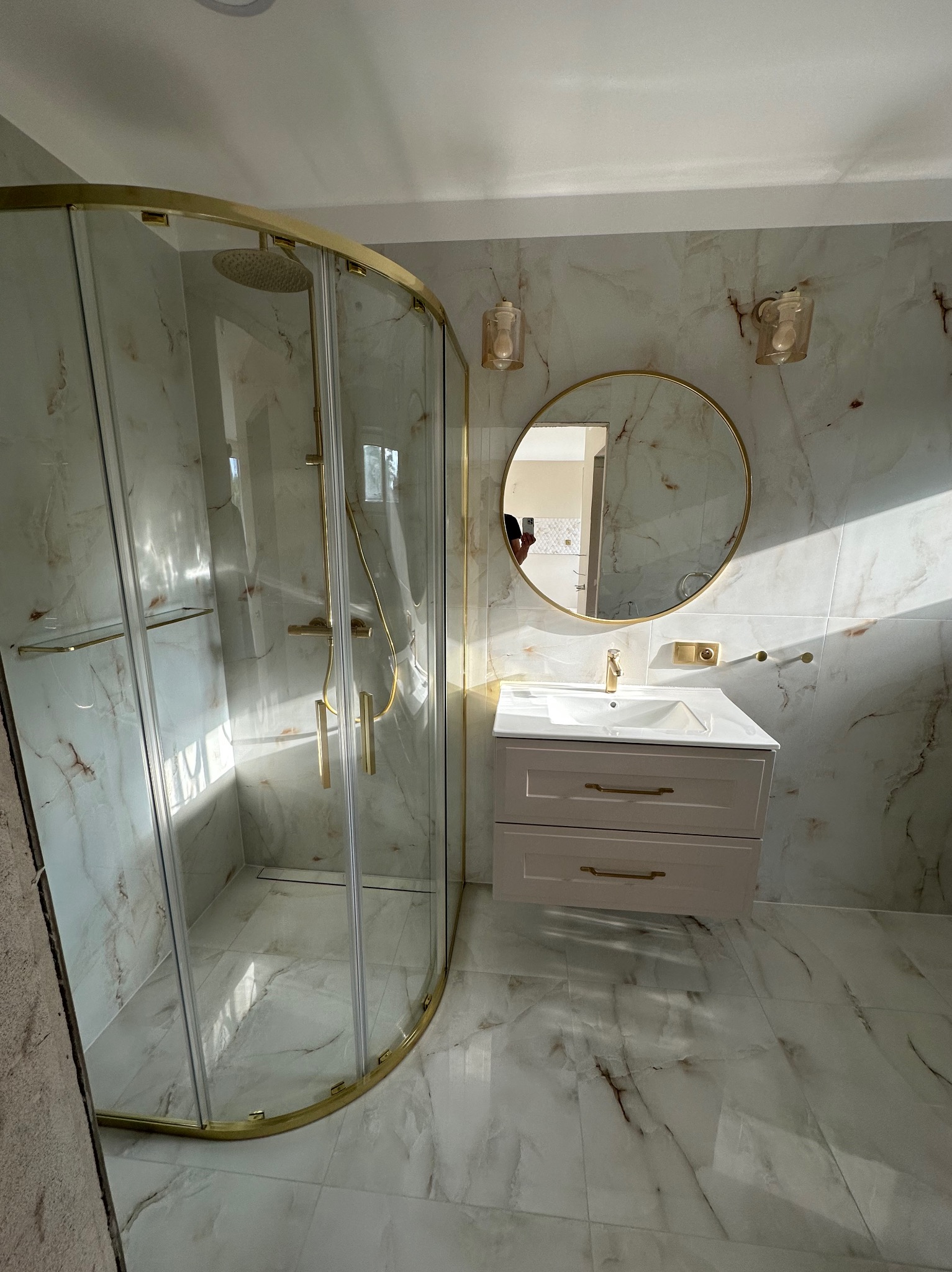 Wykończona łazienka z kabiną prysznicową w złotej ramie, okrągłym lustrem i umywalką z szafką w marmurowym wykończeniu. Nowoczesny design, jasne płytki.