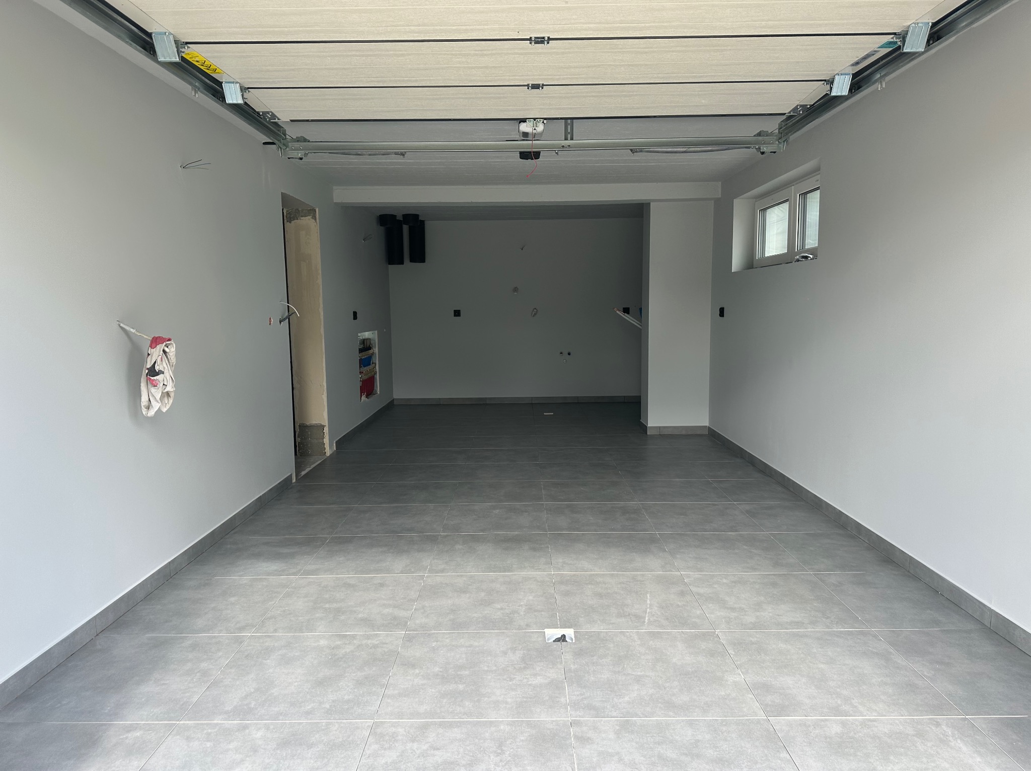 Wykończone wnętrze garażu z szarą podłogą z płytek, świeżo pomalowanymi ścianami na biało i zamontowaną bramą garażową. Widoczne instalacje.