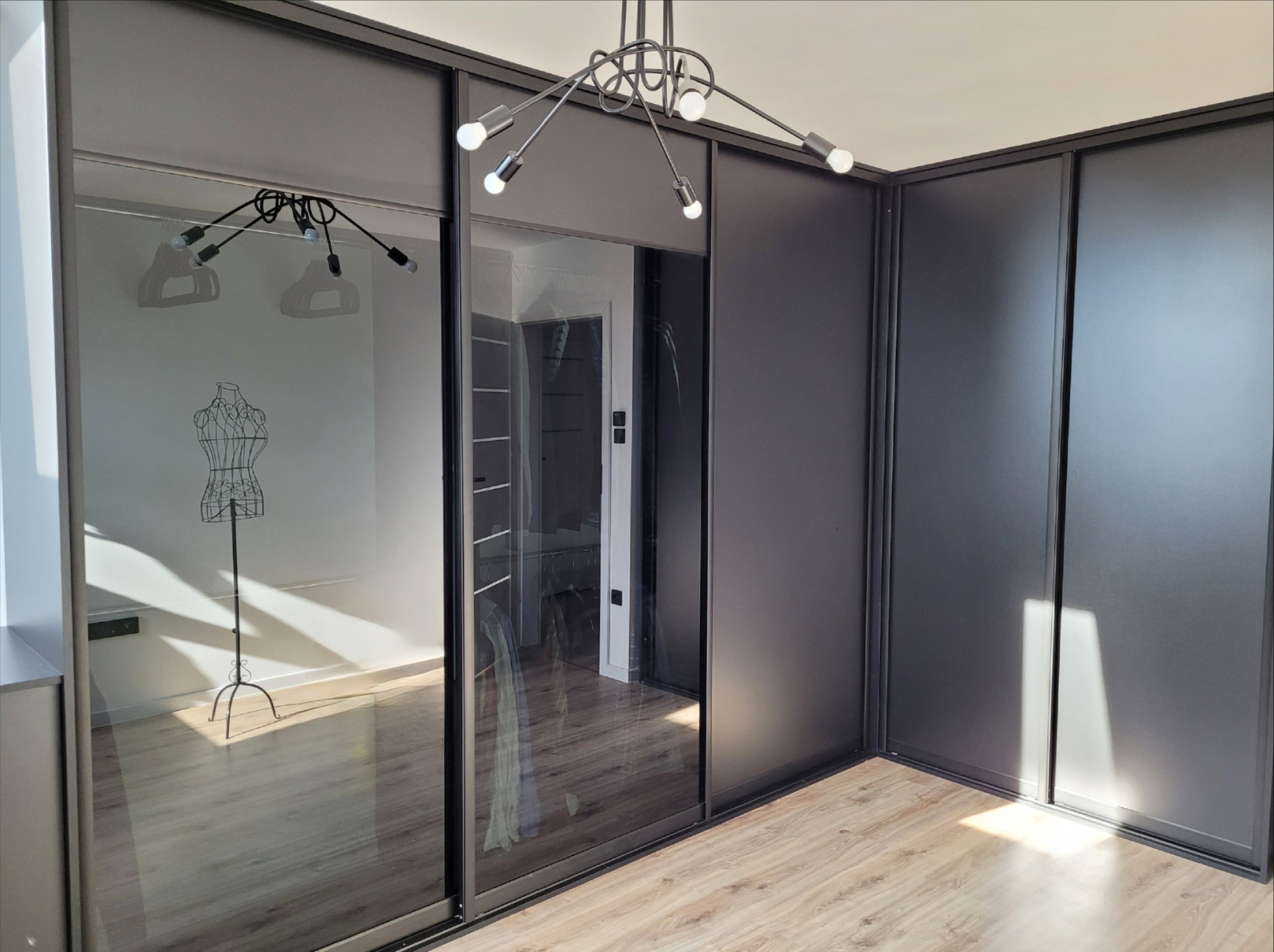 Nowoczesna szafa wnękowa z przesuwnymi drzwiami w kolorze grafitu, widoczne odbicie manekina krawieckiego i lampy sufitowej, jasna podłoga.