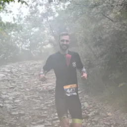 Uśmiechnięty mężczyzna z numerem startowym 443 biegnie kamienistą ścieżką w lesie, w lekkiej mgle, pokazując kciuki w górę.