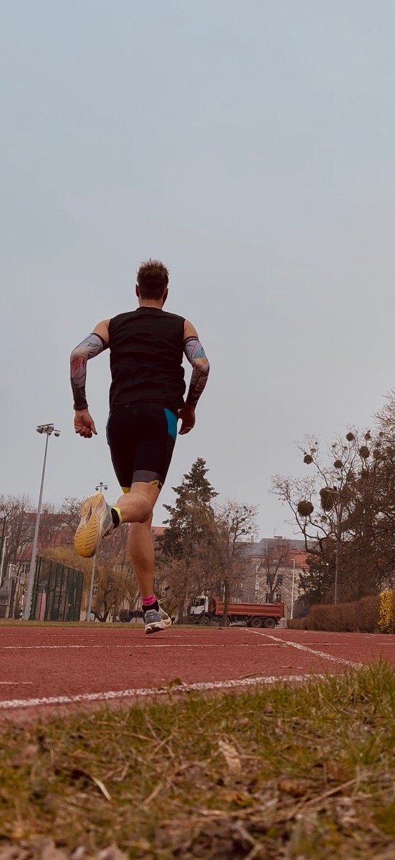 Mężczyzna z tatuażami na ramionach biegnie po czerwonej bieżni, widok z tyłu, w tle drzewa i budynki.