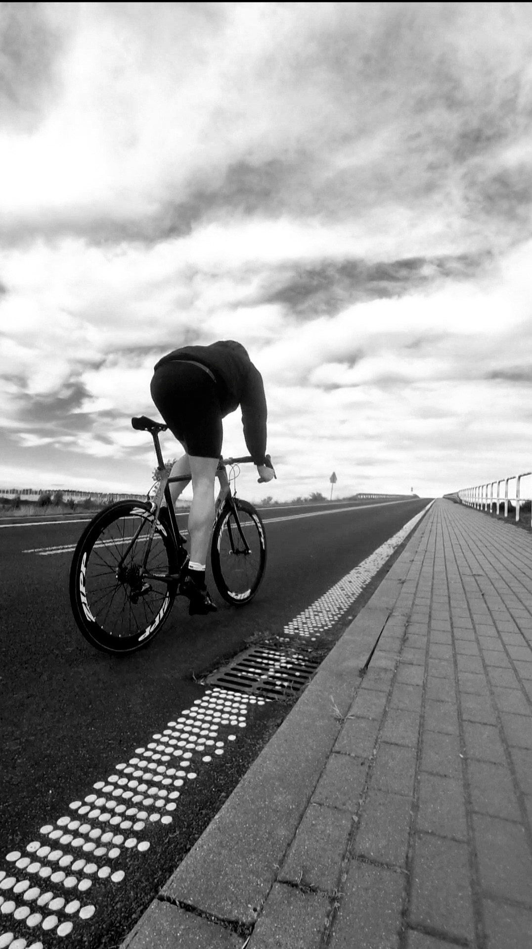 Czarno-białe zdjęcie kolarza na rowerze szosowym, jadącego wzdłuż drogi z kostki brukowej, pochmurne niebo w tle.