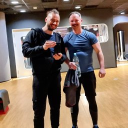 Trener Rodzinny - Dwaj uśmiechnięci mężczyźni pozują w siłowni po treningu, jeden trzyma telefon, drugi butelkę wody i ręcznik.