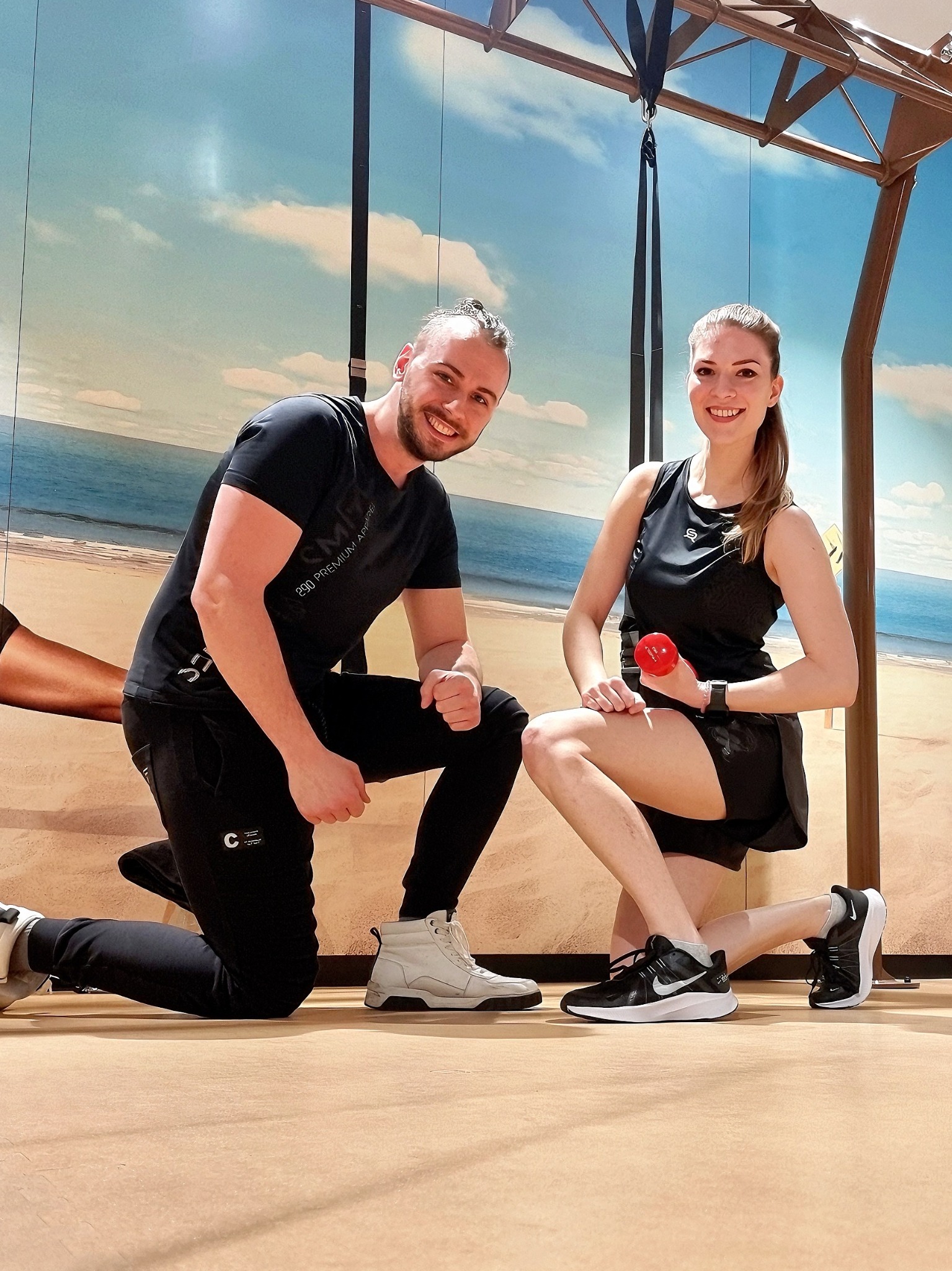 Uśmiechnięty mężczyzna w czarnym dresie i biało-szarych butach klęczy obok uśmiechniętej kobiety w czarnym sportowym stroju trzymającej czerwoną hantlę na tle dekoracji imitującej plażę i niebo.