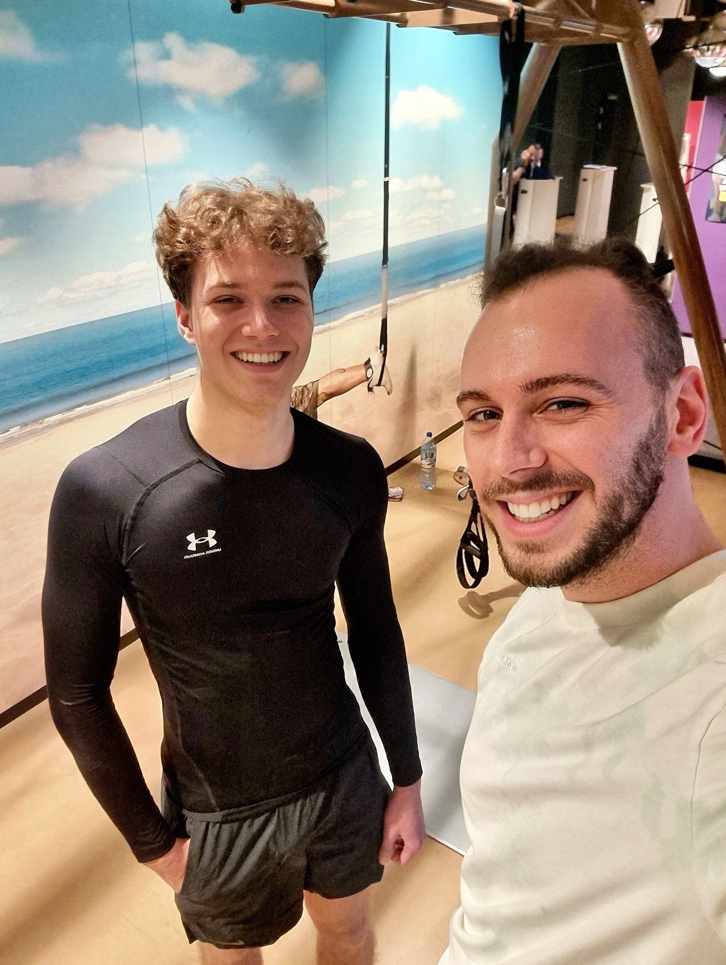 Dwaj uśmiechnięci mężczyźni w sportowych strojach pozują w sali z urządzeniami do ćwiczeń, na tle fototapety przedstawiającej plażę i morze.