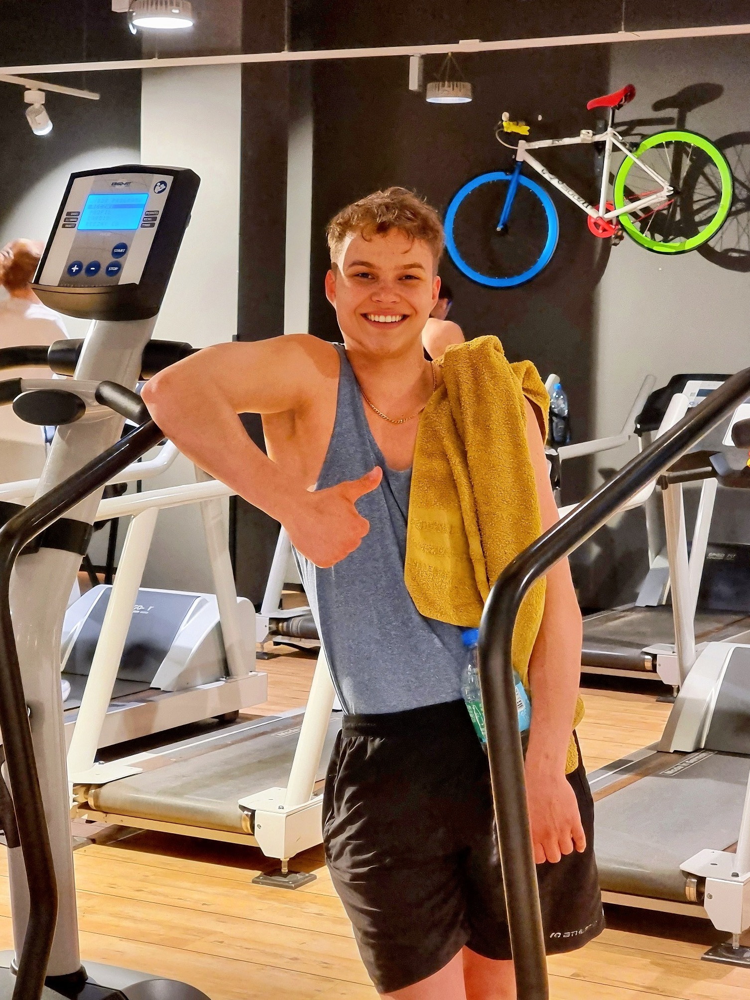 Uśmiechnięty mężczyzna z ręcznikiem na ramieniu, oparty o bieżnię w siłowni, pokazujący kciuk w górę; w tle dekoracja ścienna w kształcie roweru z neonowymi kołami.