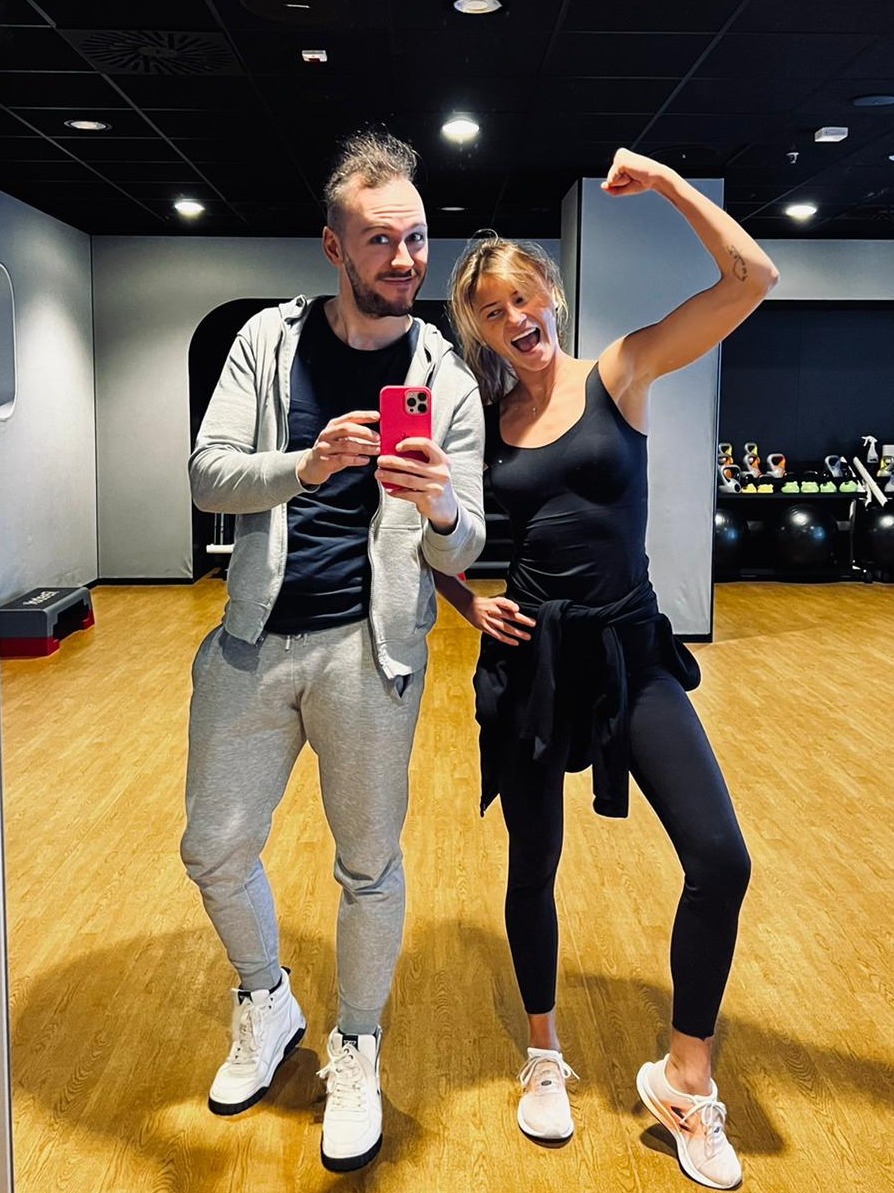 Dwie uśmiechnięte osoby w strojach sportowych pozują do selfie w siłowni, kobieta z uniesioną ręką prezentuje biceps, w tle widoczne sprzęty do ćwiczeń.