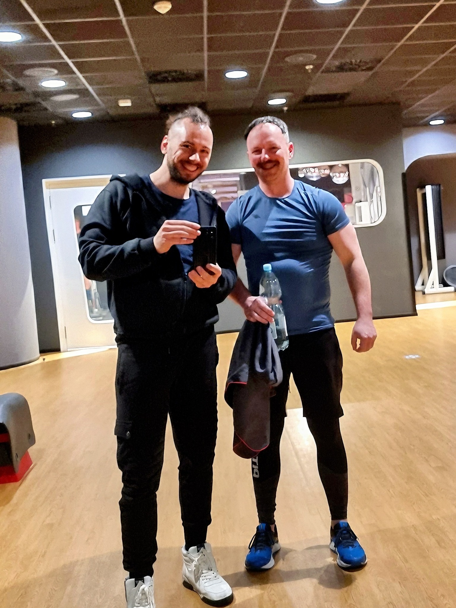 Dwaj uśmiechnięci mężczyźni pozują w siłowni po treningu, jeden trzyma telefon, drugi butelkę wody i ręcznik.