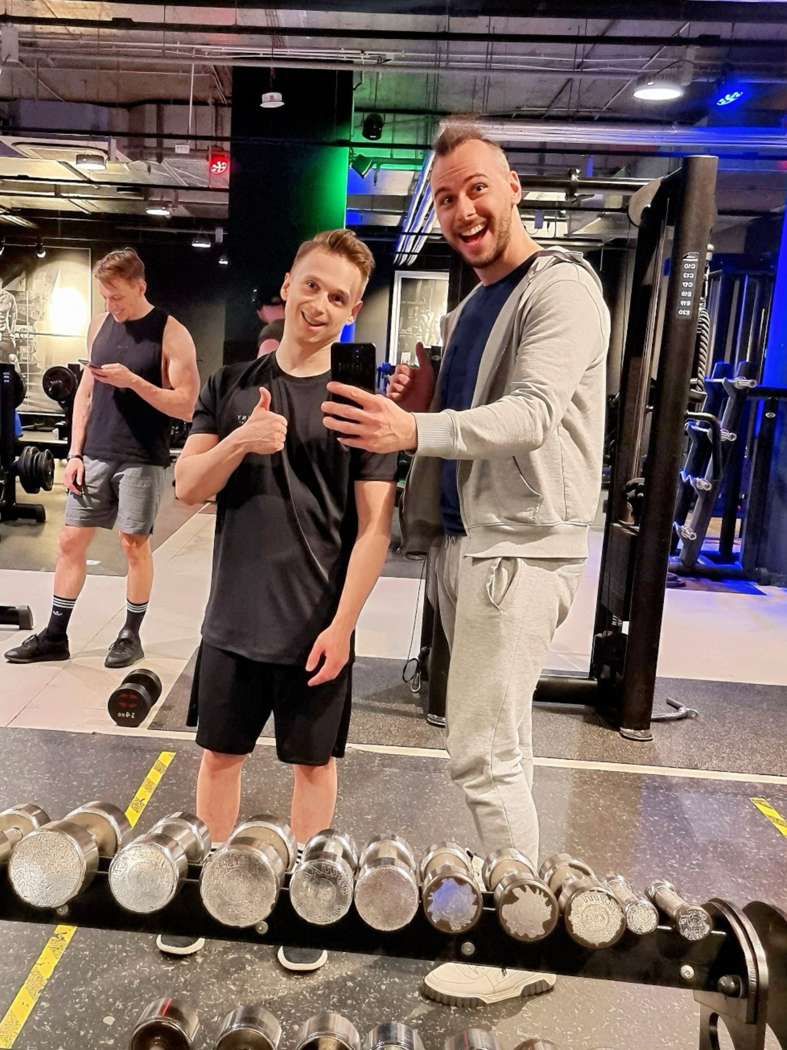 Dwaj uśmiechnięci mężczyźni pozują do selfie w siłowni, jeden w sportowym dresie, drugi w czarnym stroju sportowym, obok stojak z hantlami, w tle ćwiczący mężczyzna i maszyny do ćwiczeń.