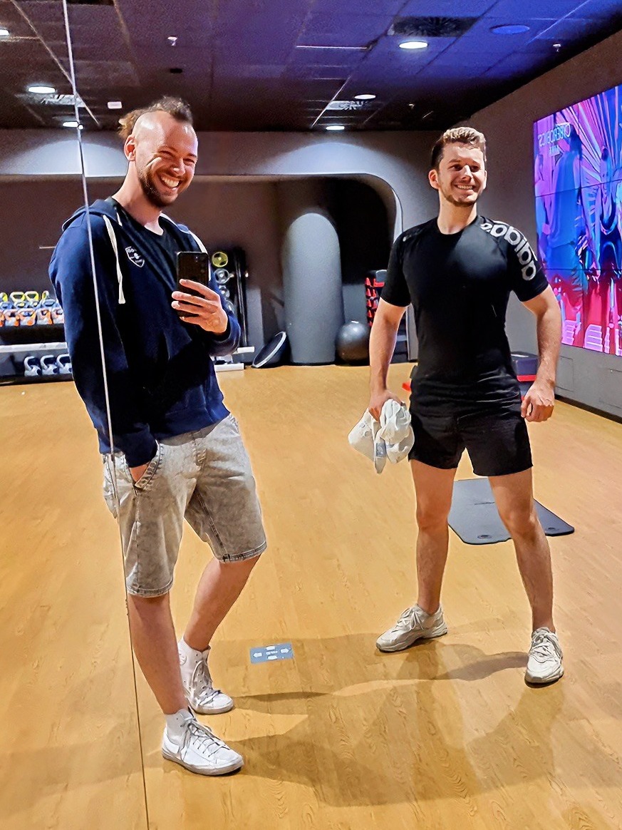 Dwóch uśmiechniętych mężczyzn pozuje do selfie w siłowni, widoczny sprzęt do ćwiczeń i mata do jogi