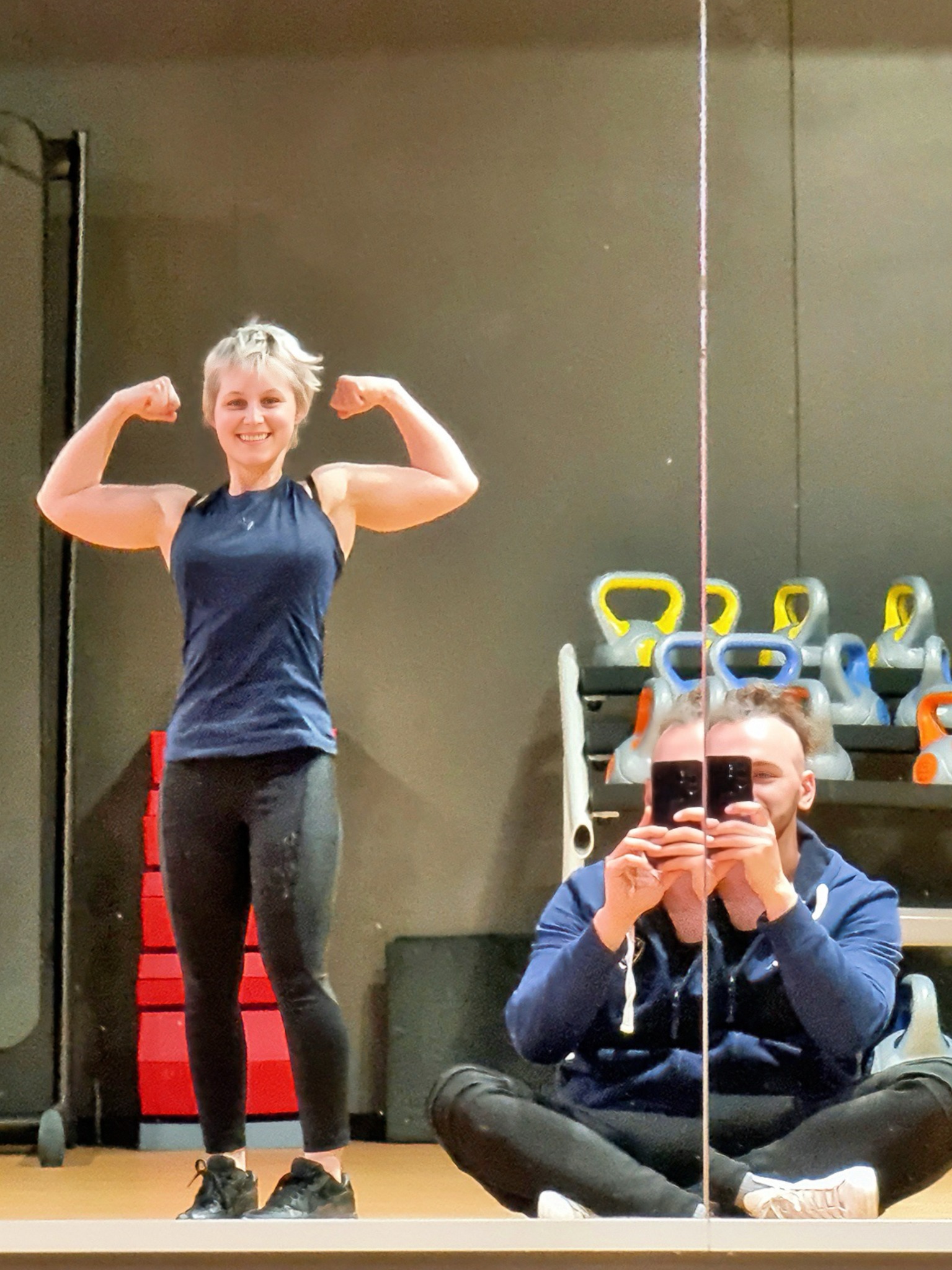 Uśmiechnięta kobieta w sportowym stroju, prezentująca napięte bicepsy, sfotografowana w lustrze siłowni; w odbiciu mężczyzna robiący jej zdjęcie telefonem, w tle kettlebells i drabinka gimnastyczna.