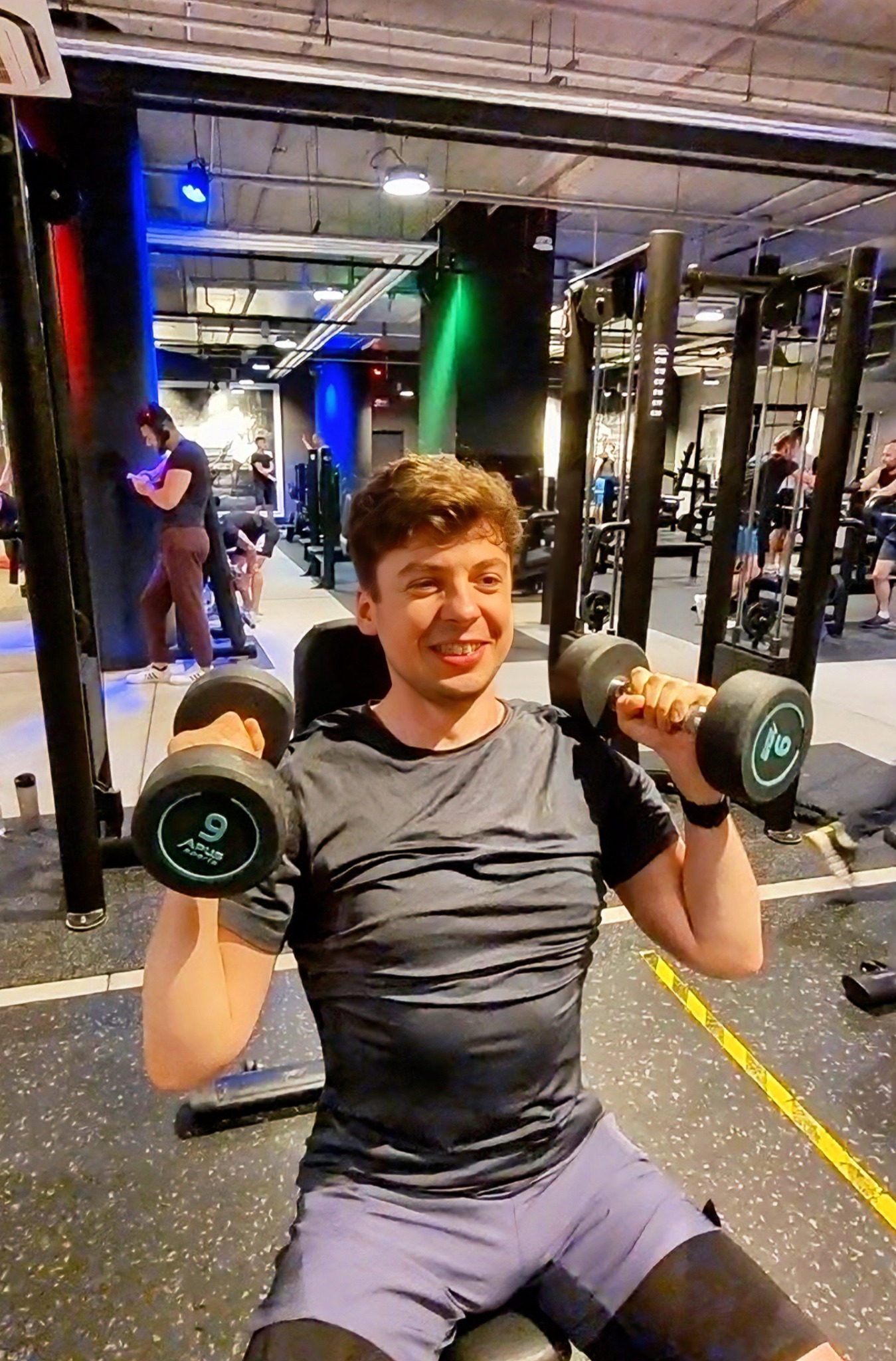 Uśmiechnięty mężczyzna siedzi na ławce w siłowni, podnosząc dwie ciężarki z numerem 9, w tle widoczne inne osoby i sprzęt do ćwiczeń.
