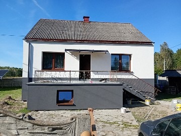 Dwukondygnacyjny dom z nową, dwukolorową elewacją: białą na górze i szarą na dole. Widoczne schody wejściowe i balkon z czarną balustradą. Dach pokryty szarą dachówką.