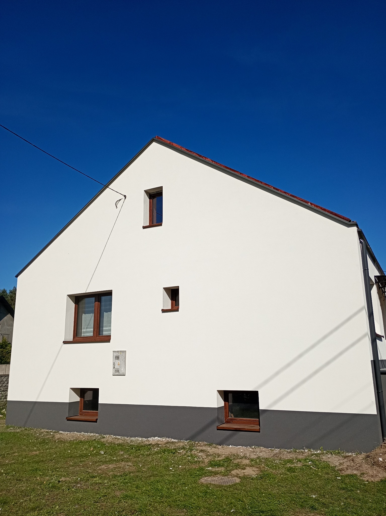 Biała elewacja domu z brązowymi oknami na tle błękitnego nieba. Widoczny fragment dachu i instalacja elektryczna. Dolna część elewacji pomalowana na szaro.