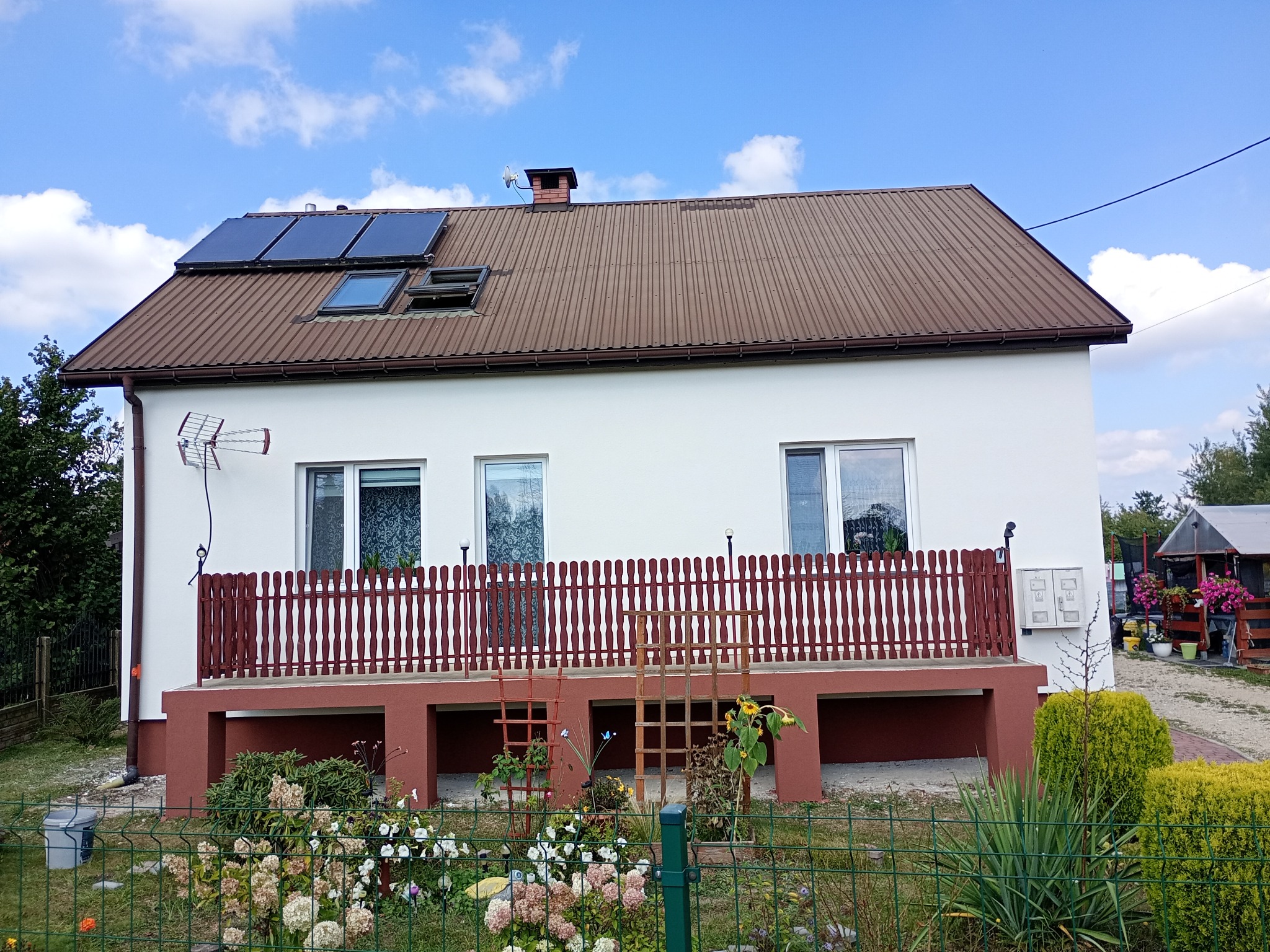 Dwukondygnacyjny dom z brązowym dachem, panelami słonecznymi i balkonem z drewnianą balustradą. Biała elewacja, zadbany ogród z kwiatami i zielonym ogrodzeniem.